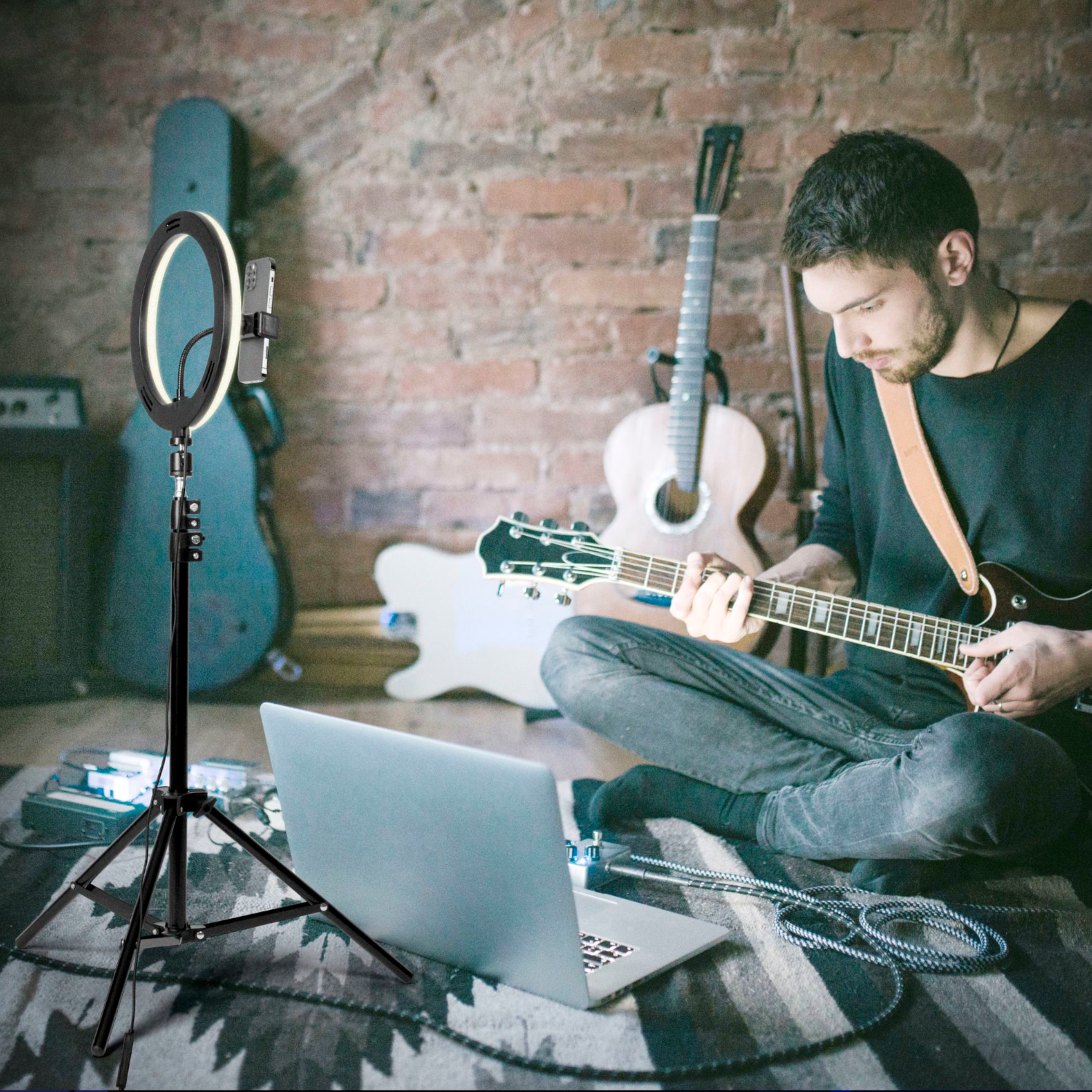A man playing guitar in a room with a ring light and a laptop. Guitars and a brick wall background.