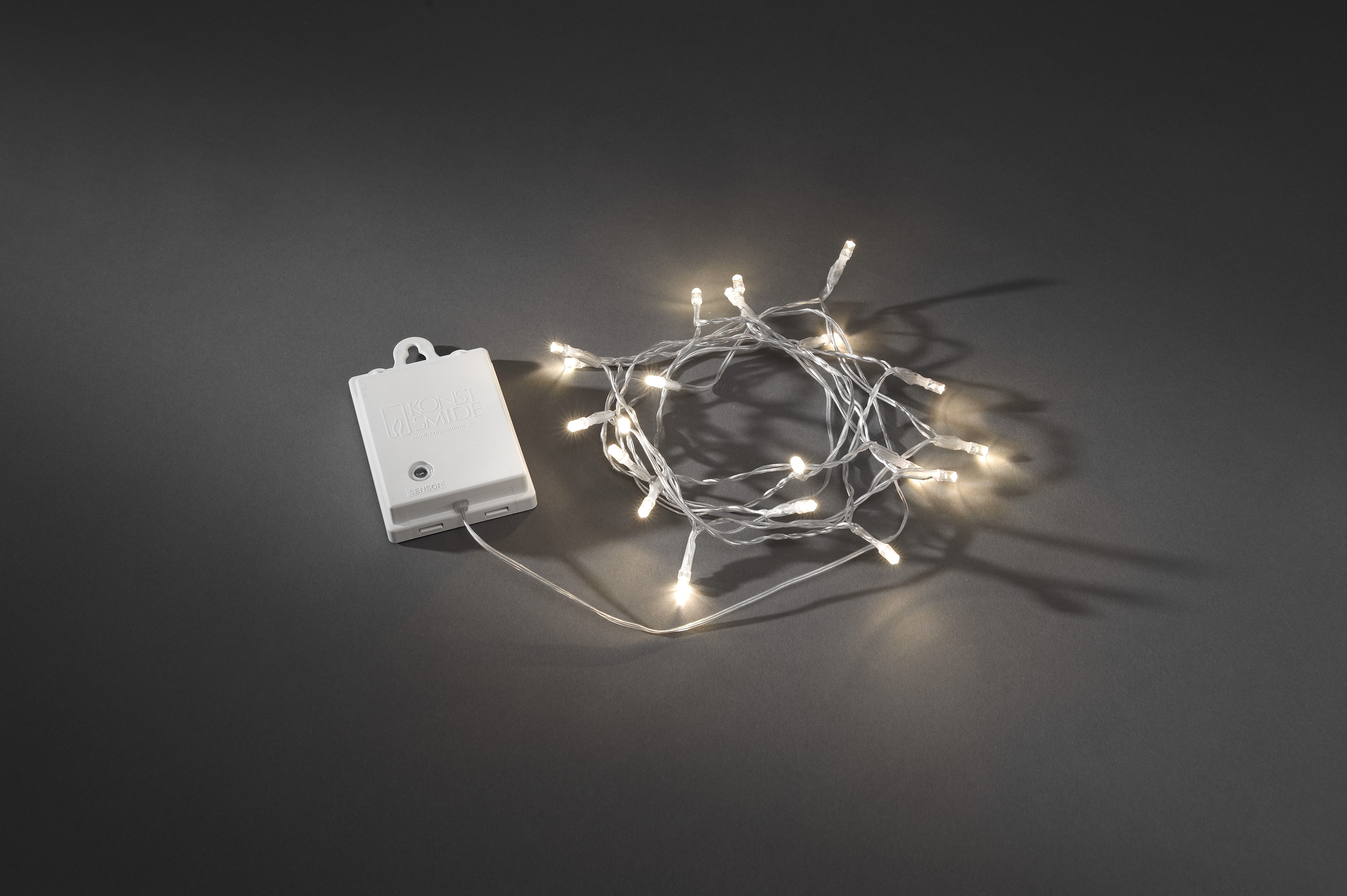 Eine Lichterkette mit kleinen weißen Lichtern und einem Akkupack auf dunkelgrauer Oberfläche.