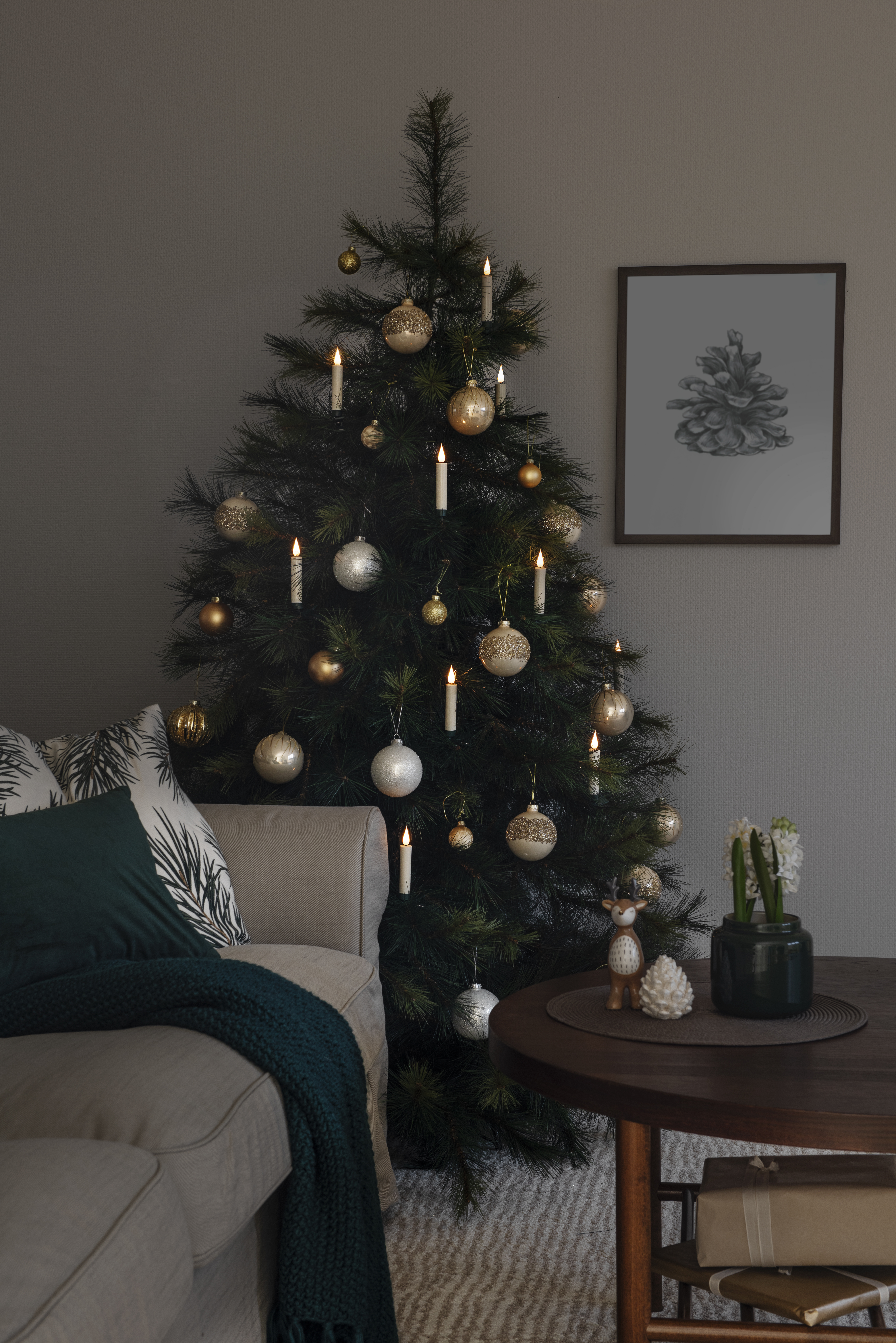 Ein geschmückter Weihnachtsbaum steht neben einem Sofa mit einem grünen Überwurf. Ein Tisch hat eine Vase, Geschenke und eine Hirschfigur.