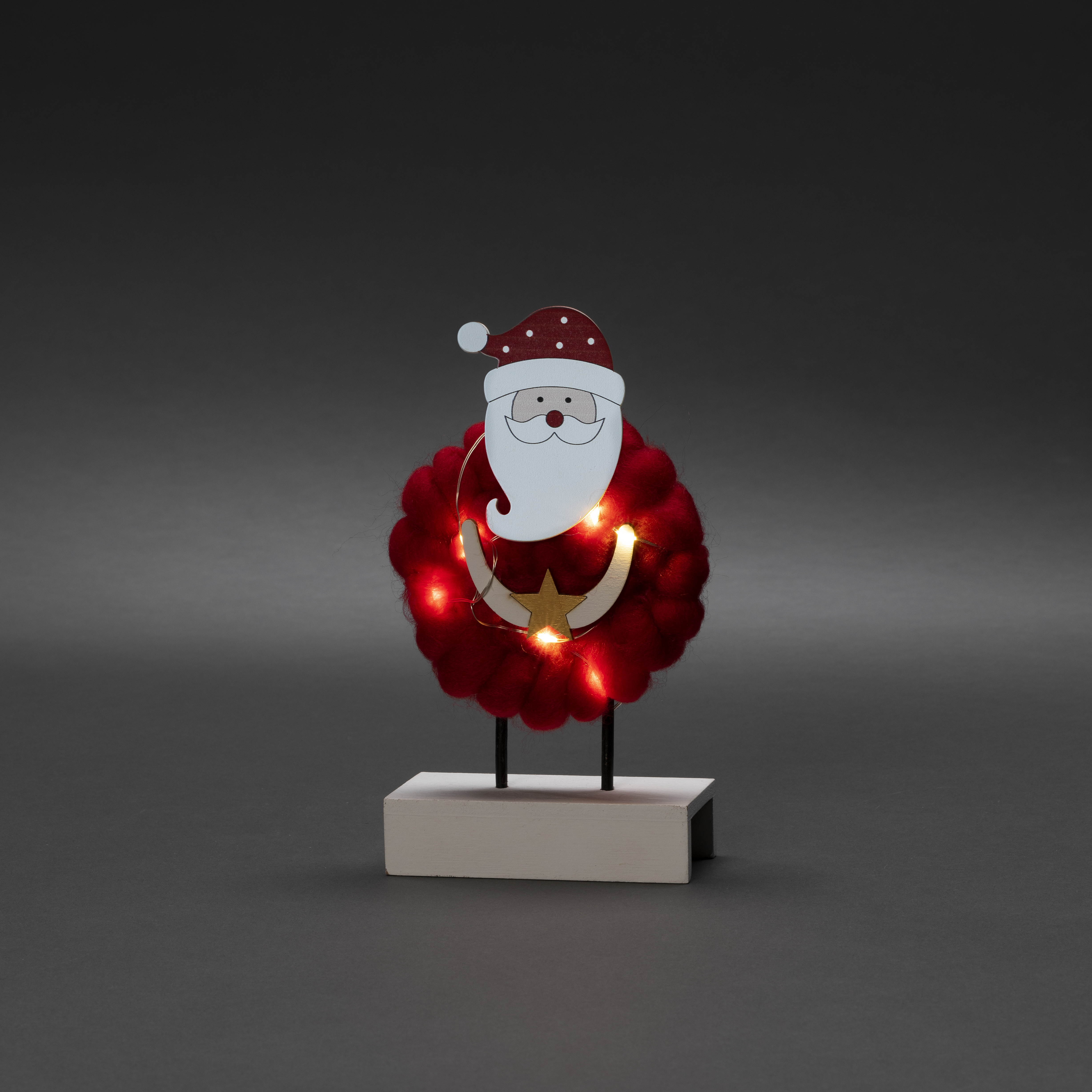 Weihnachtliche Santa-Dekoration aus Holz mit rotem Bart und Hut, umgeben von Lichtern, auf weißem Sockel.