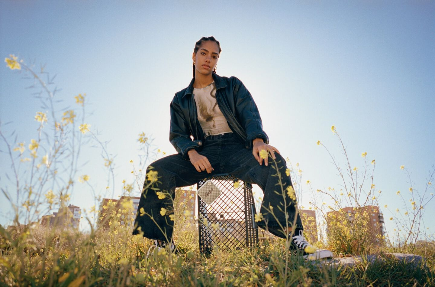 Frau sitzt auf einer Kiste in einem Feld mit gelben Blumen, trägt Jeansjacke und -hose. Blauer Himmel im Hintergrund.