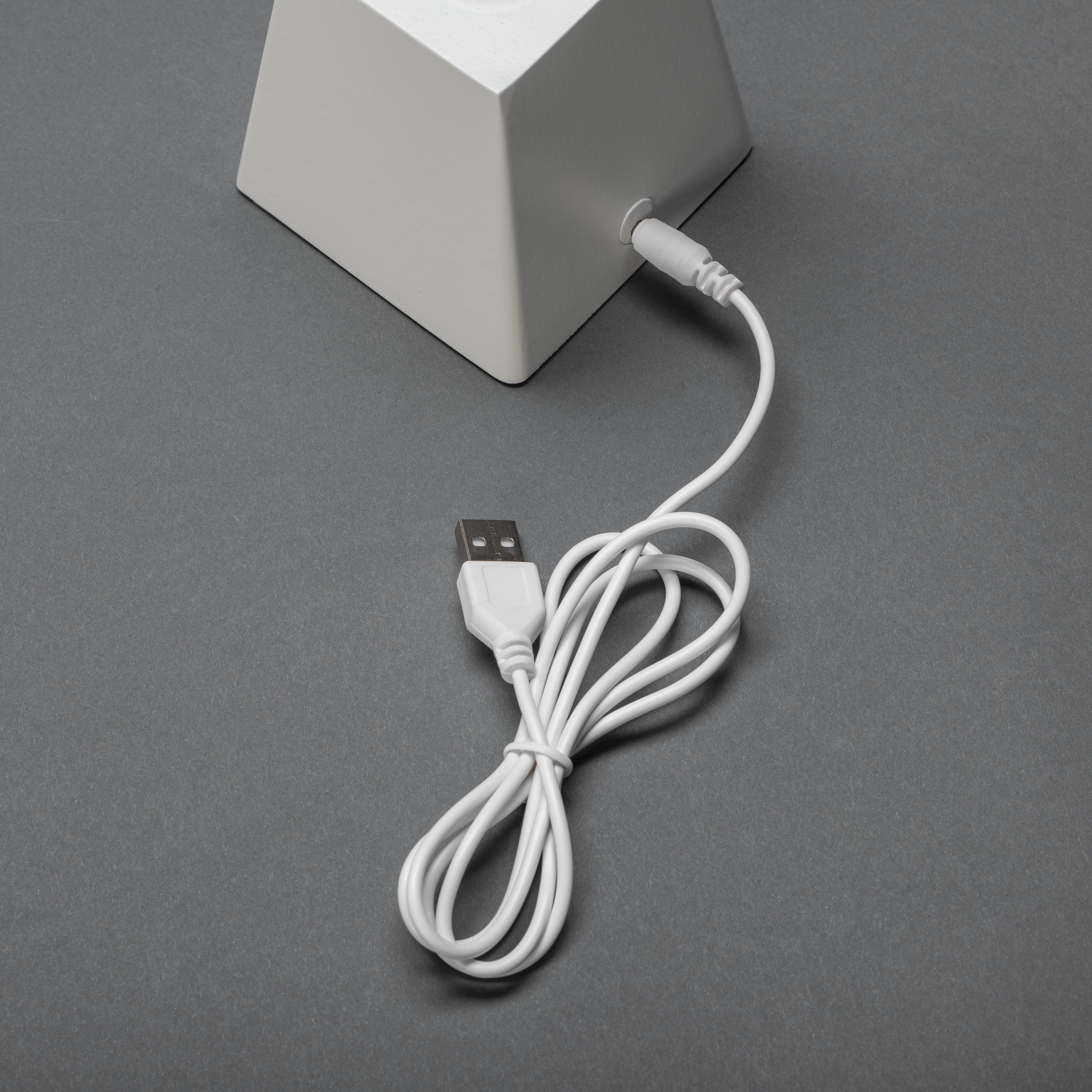 Ein weißer Sockel mit einem weißen USB-Kabel, das auf einer grauen Oberfläche liegt. Das Kabel ist geschlungen.