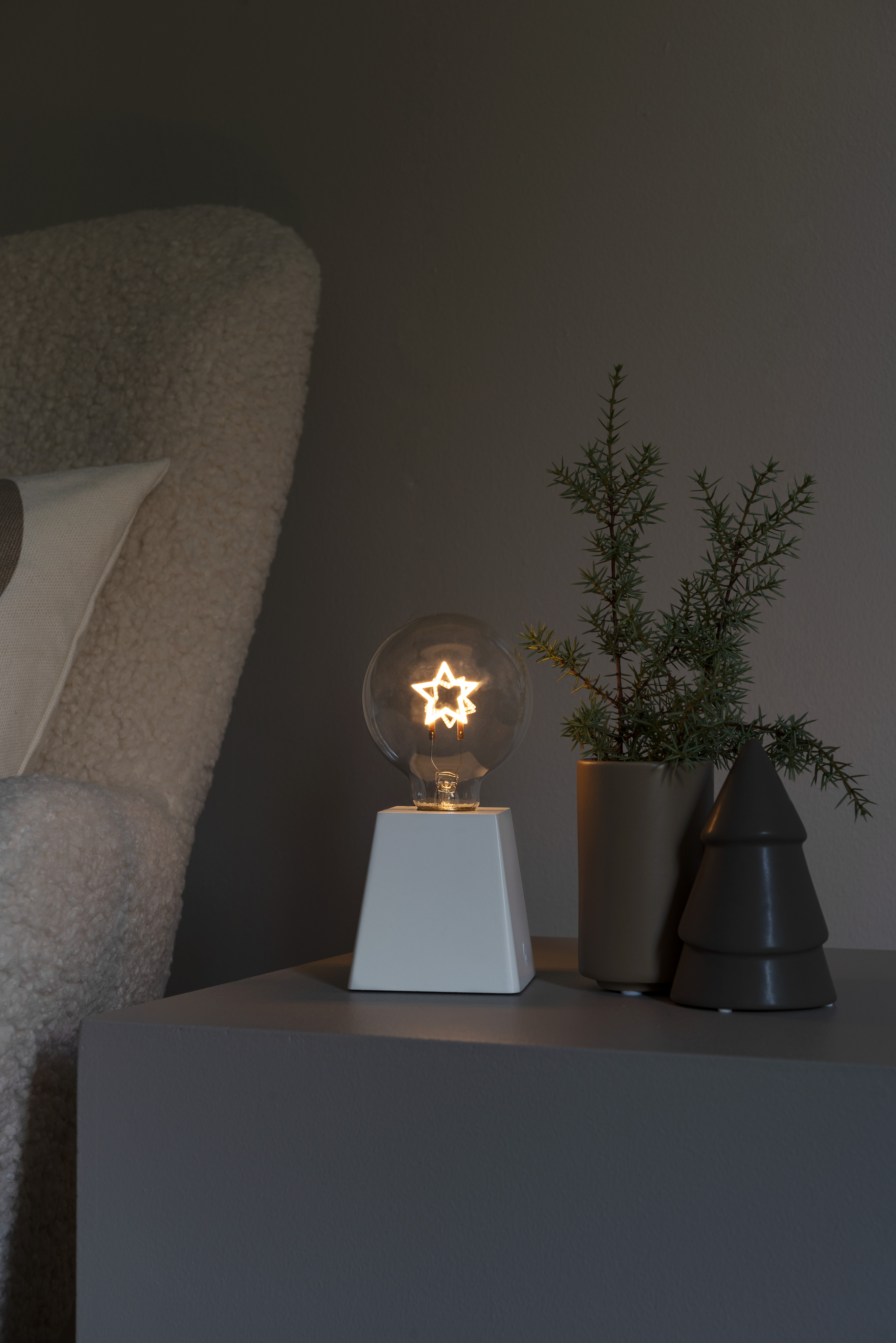 Eine Lampe mit sternförmiger Glühbirne und einem Weihnachtsbaum daneben. Neutrale Töne.