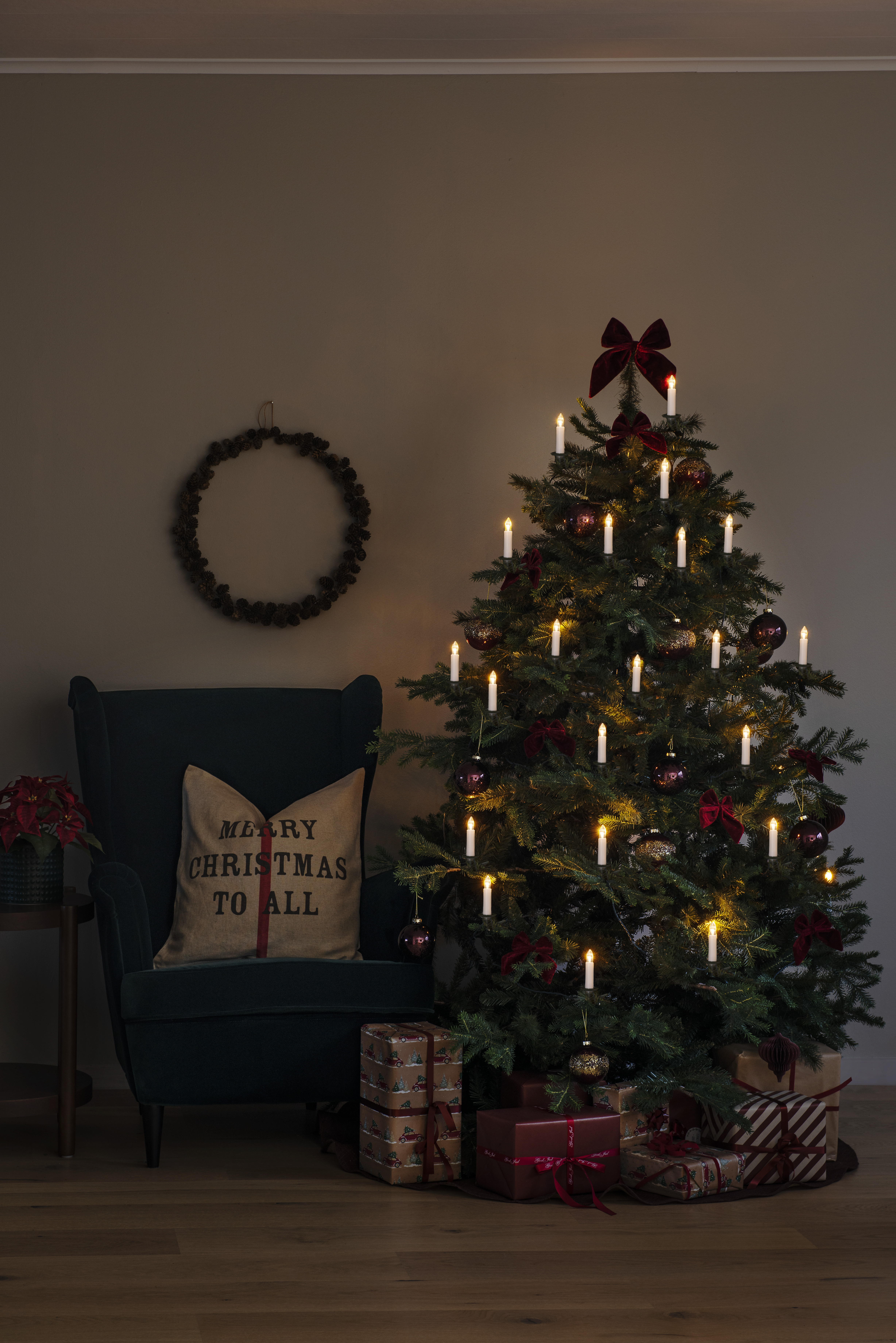 Weihnachtsbaum mit Lichtern, Ornamenten und Geschenken. Es gibt ein Merry Christmas Kissen und einen Kranz.
