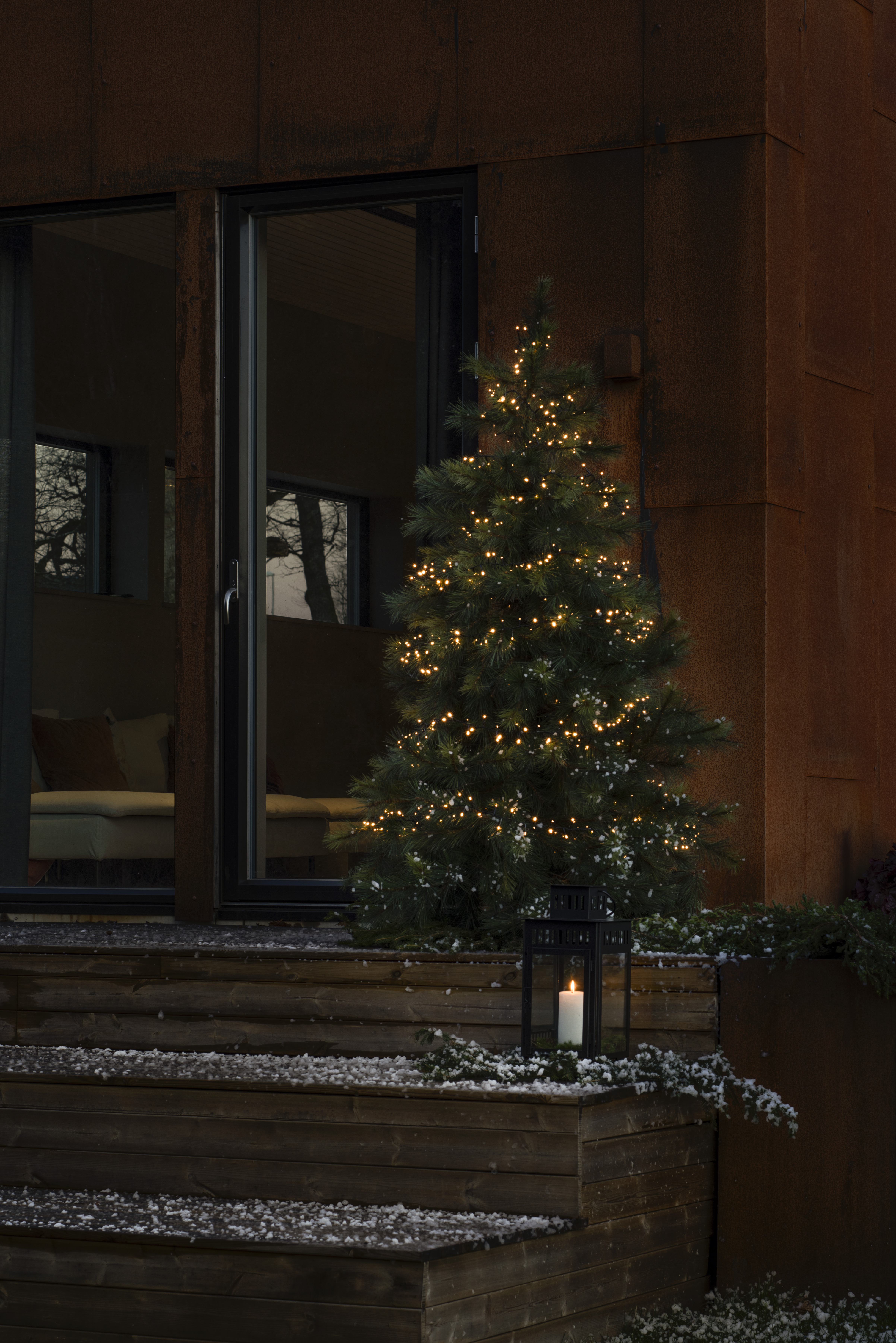 Ein Weihnachtsbaum mit Lichtern steht vor einem Haus. Eine brennende Kerze steht in einer Laterne auf den Stufen.