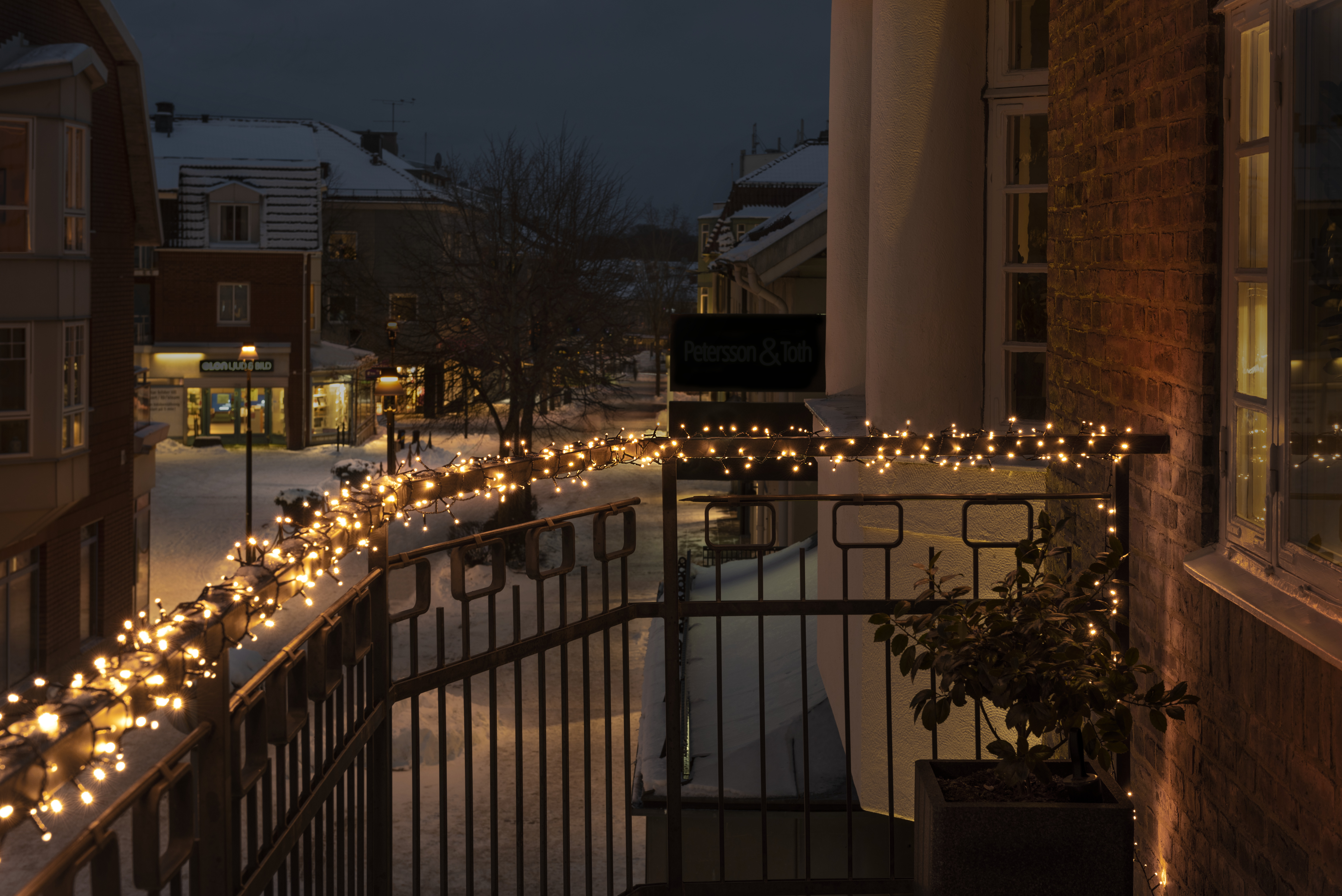 Nachtszenen mit Gebäuden, Schnee und Lichterketten. Ein Balkon mit Lichtern und einer Pflanze. Gebäude im Hintergrund.