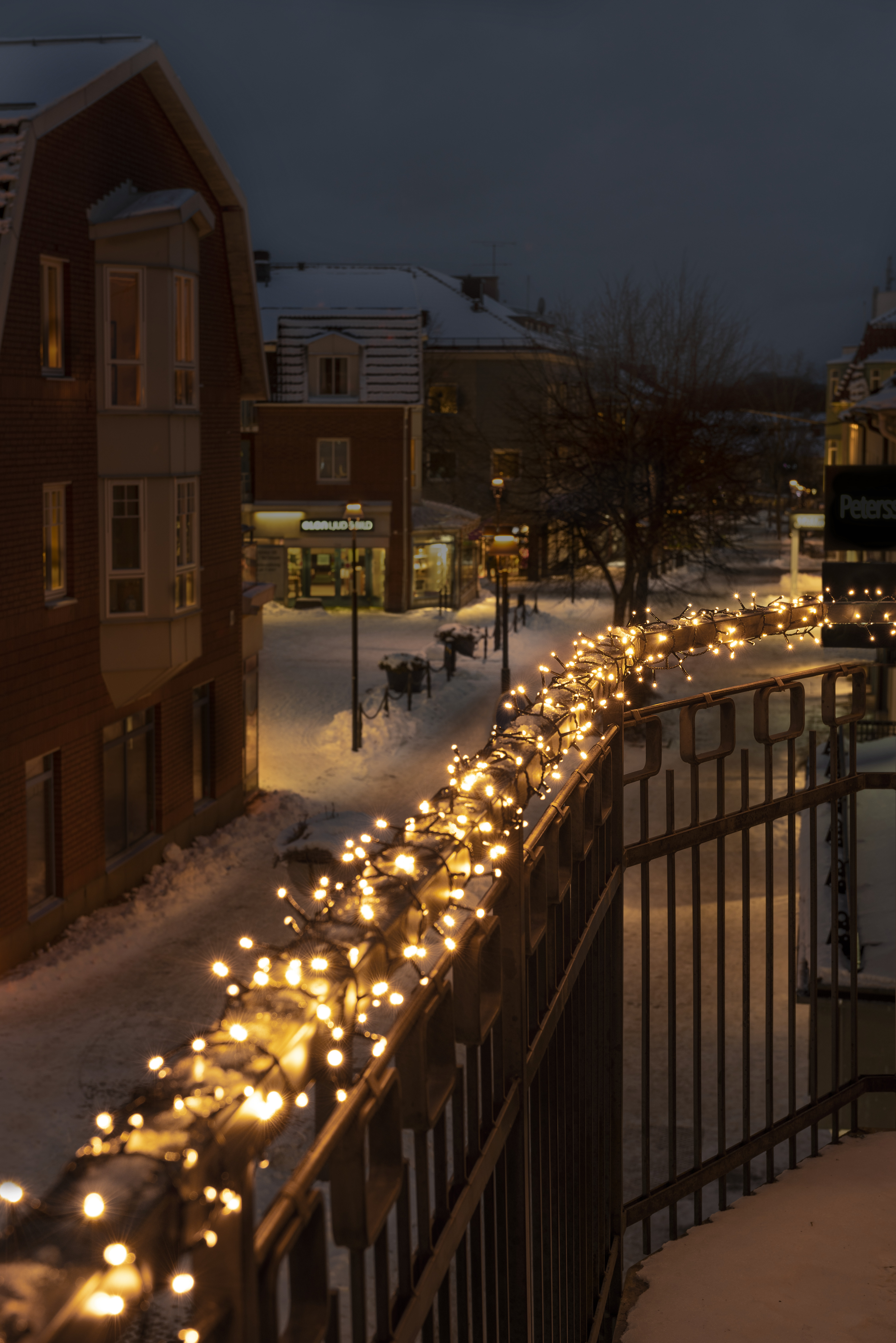 Winterliche Straßenszene in der Dämmerung. Lichterketten schmücken ein Geländer. Gebäude und schneebedeckte Straßen sind sichtbar.