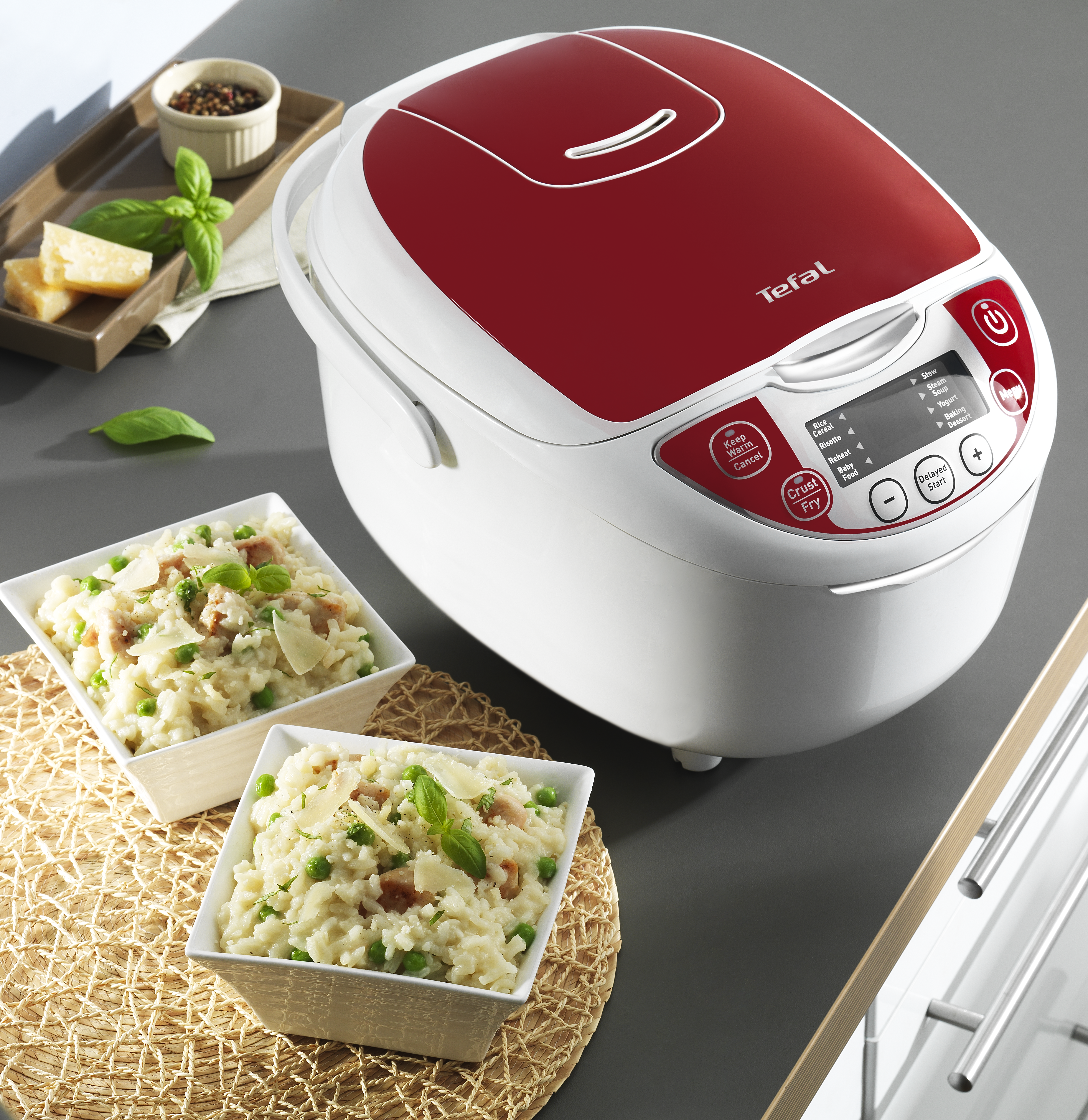 Een witte en rode Tefal rijstkoker staat naast twee kommen risotto. Ingrediënten als kaas en basilicum zijn ook aanwezig.