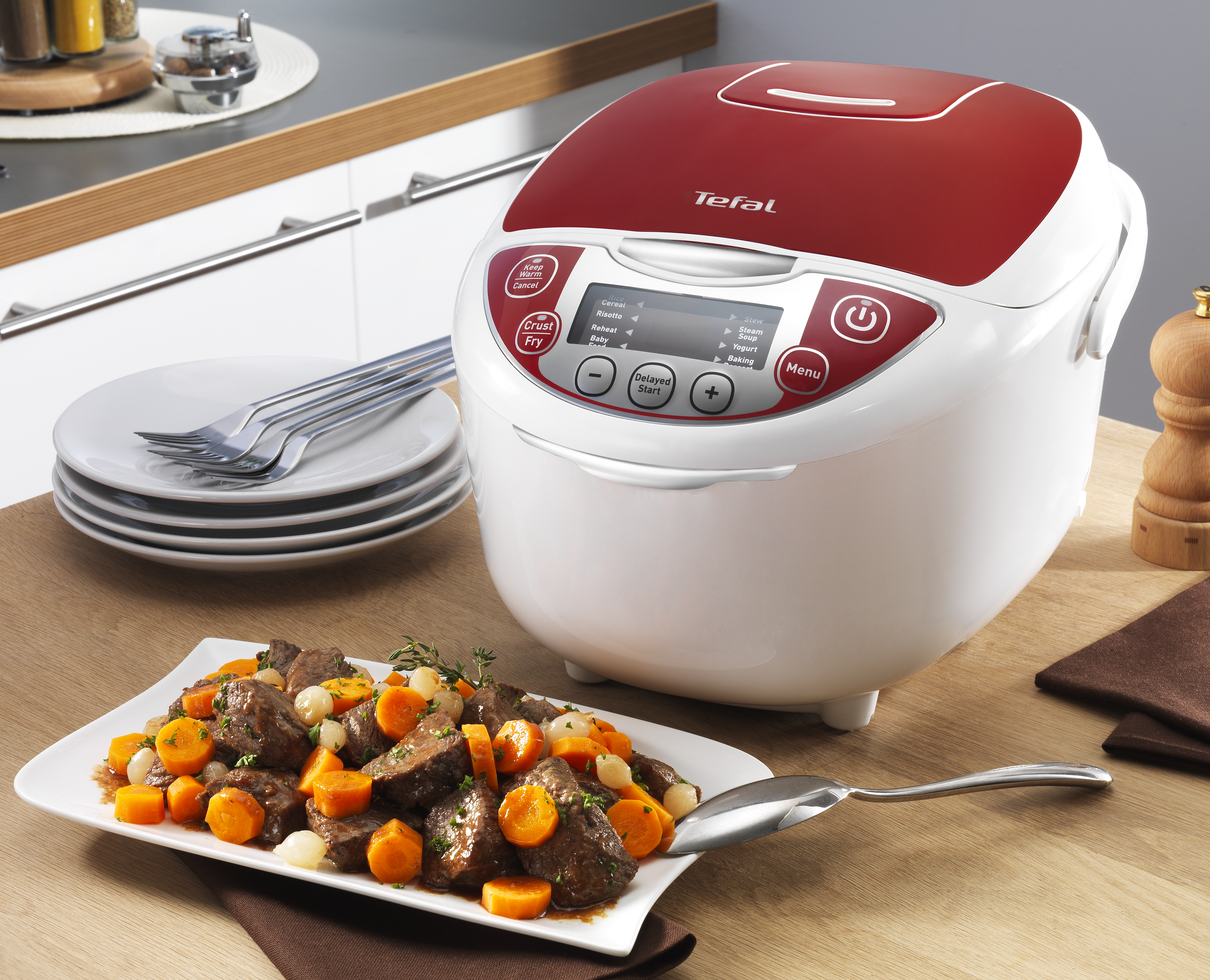 Een Tefal rijstkoker, een bord eten en borden op een houten tafel. De koker heeft een rood deksel en een witte behuizing.