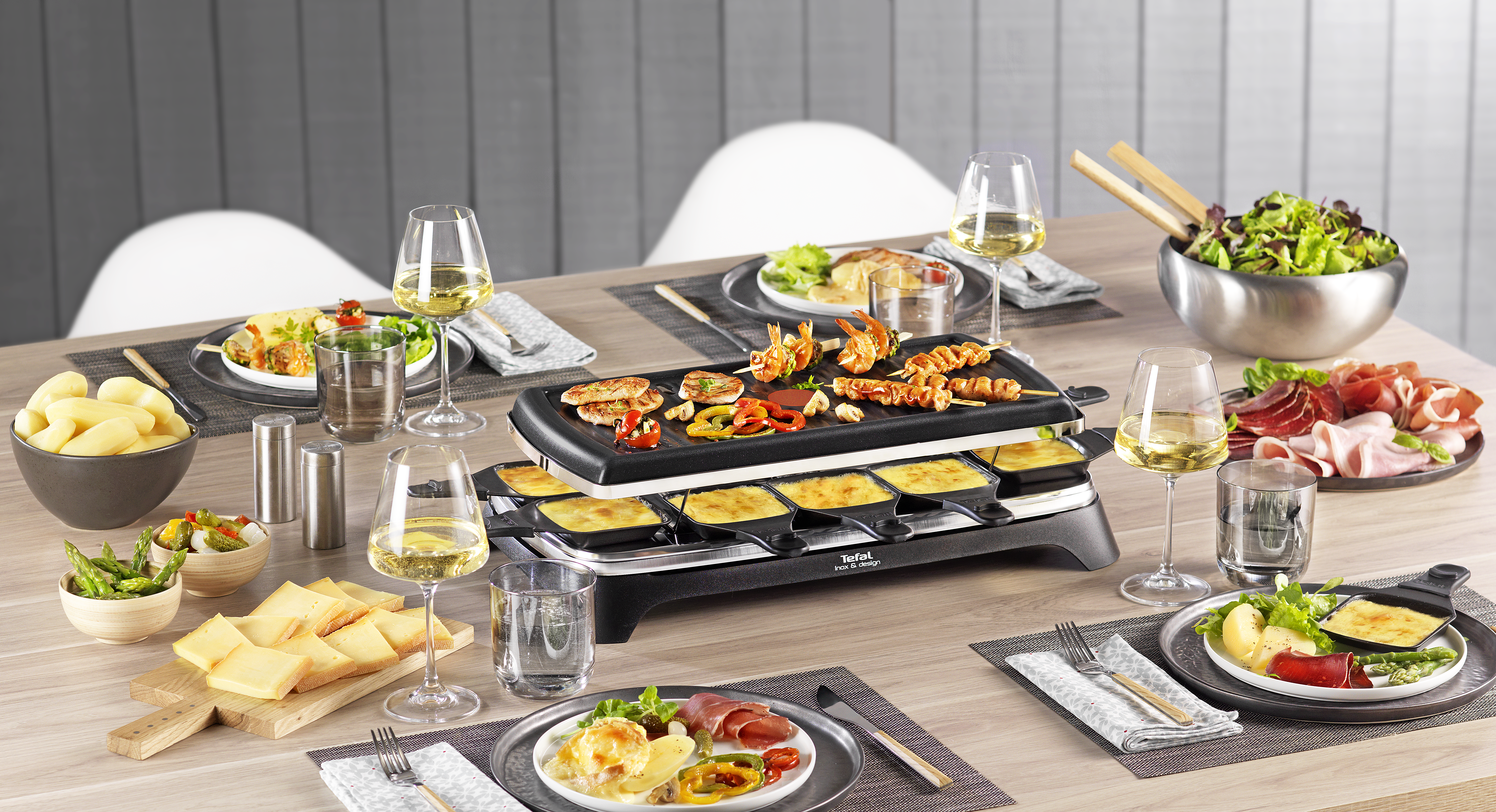 A table set for raclette with grilled food, cheese, wine, and various side dishes. A raclette grill is in the center.