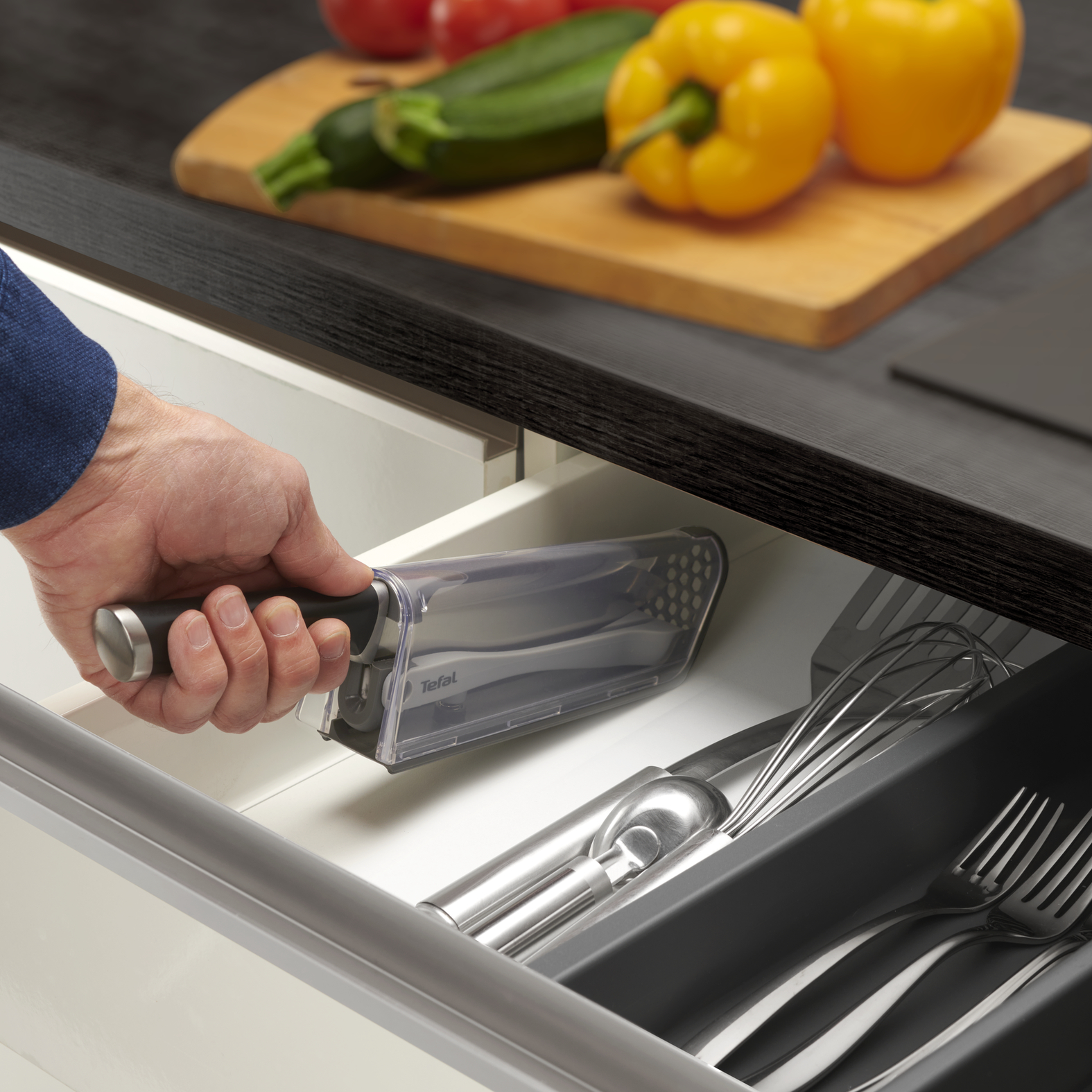Una mano sosteniendo un cuchillo en un cajón con cubiertos y verduras detrás.
