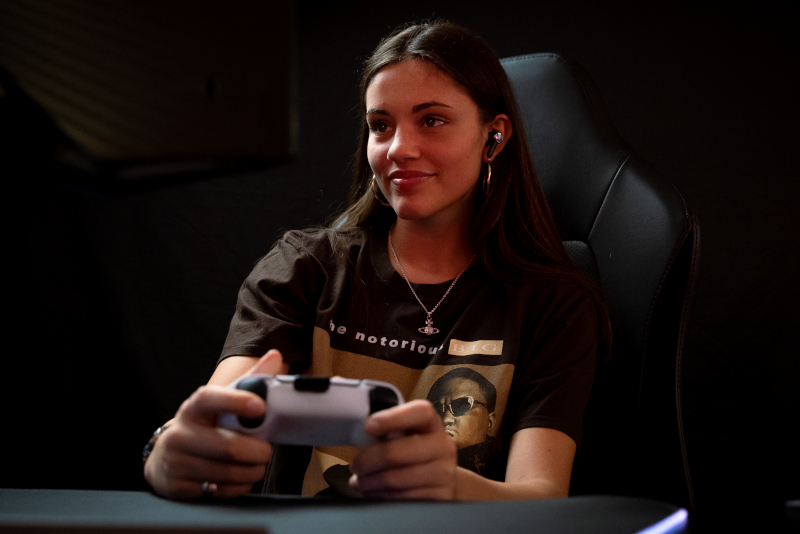 Frau spielt Videospiel, hält Controller, trägt Ohrhörer, dunkler Raum.