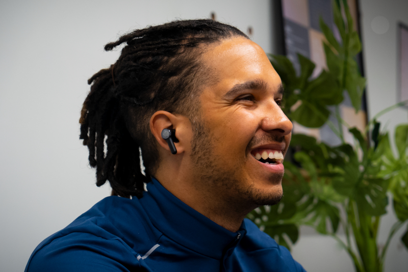 Mann mit Dreadlocks lächelt, trägt ein blaues Hemd, Ohrhörer, schaut zur Seite.