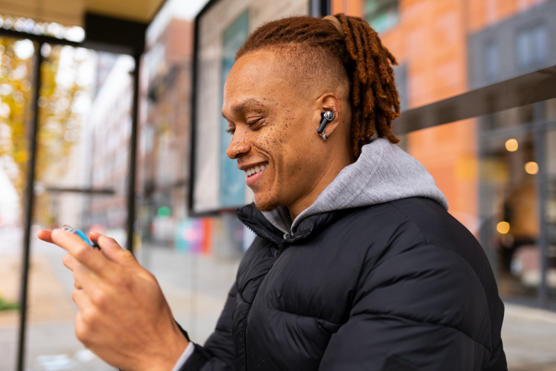 Ein Mann mit Dreadlocks lächelt, während er auf ein Telefon schaut. Er trägt eine schwarze Jacke und hat Ohrhörer drin.