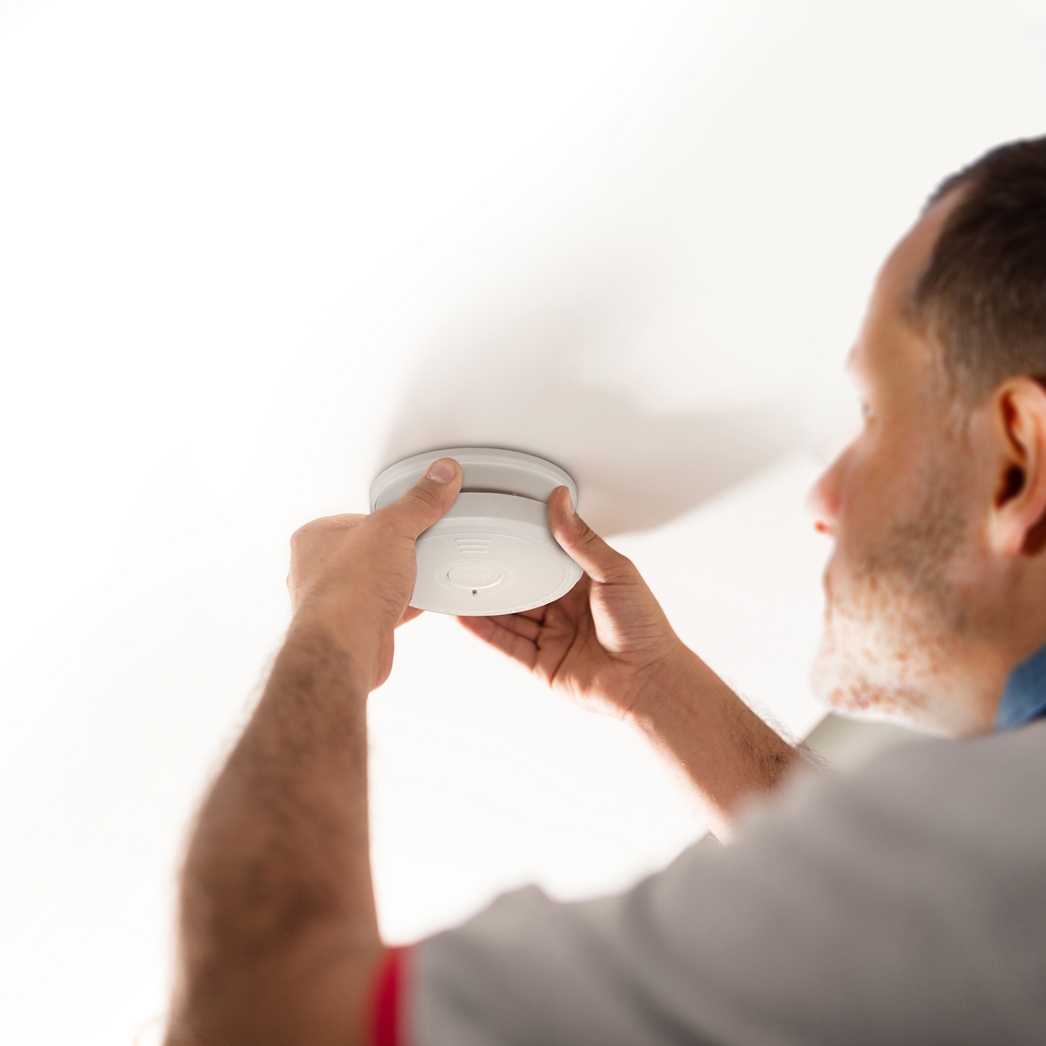 Een man installeert een witte rookmelder aan een wit plafond. Focus op de handen en het apparaat.