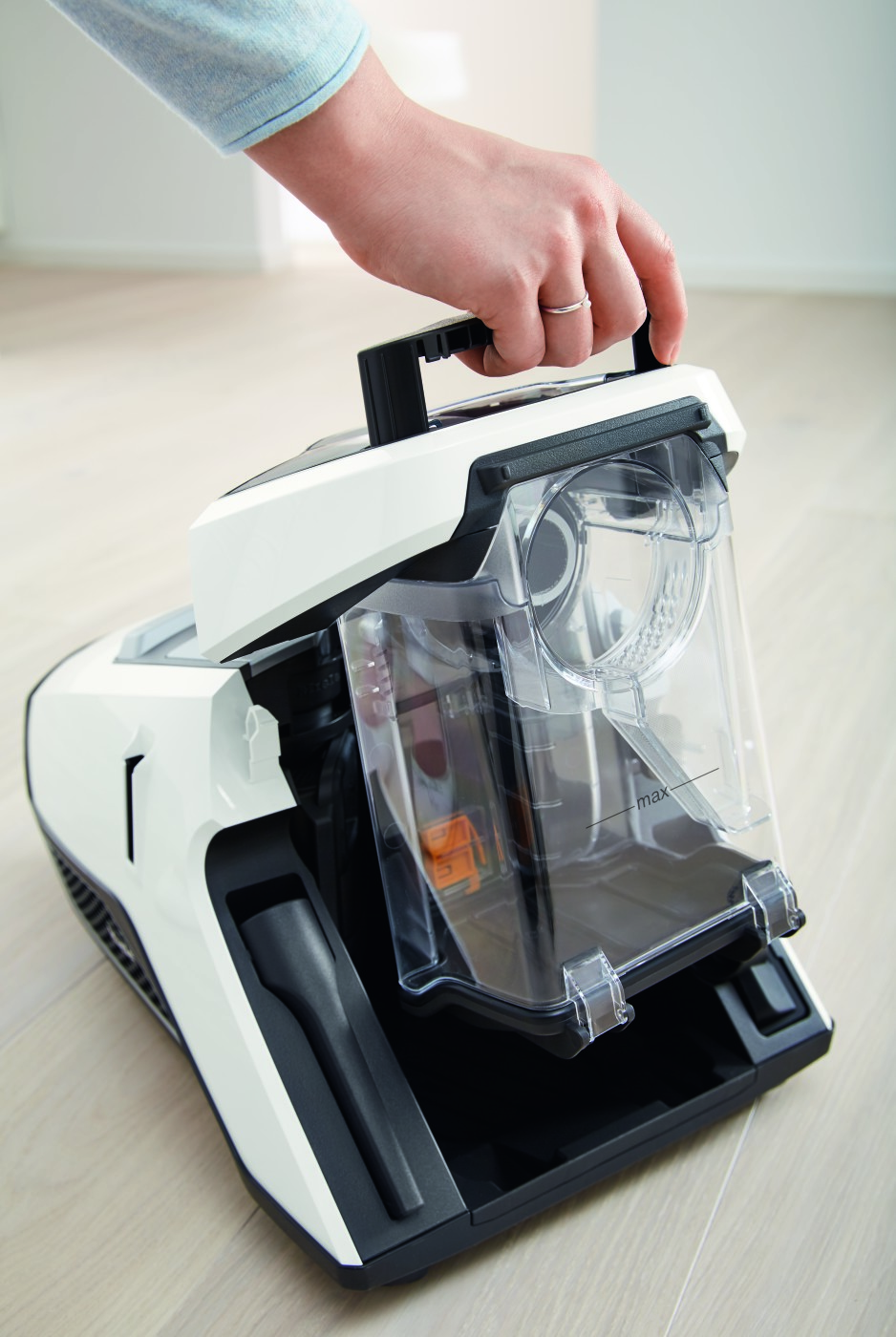 A hand holding the handle of a white and black vacuum cleaner with a transparent dust container.