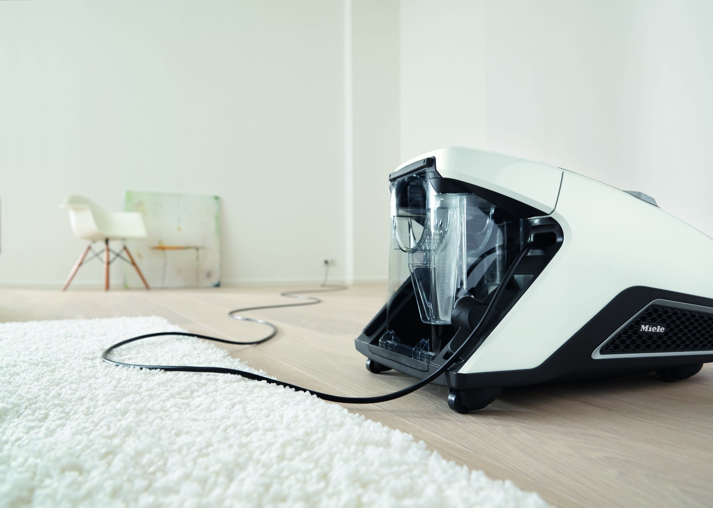 White vacuum cleaner on a white carpet, cord stretched across the floor. Background has a chair and wall.