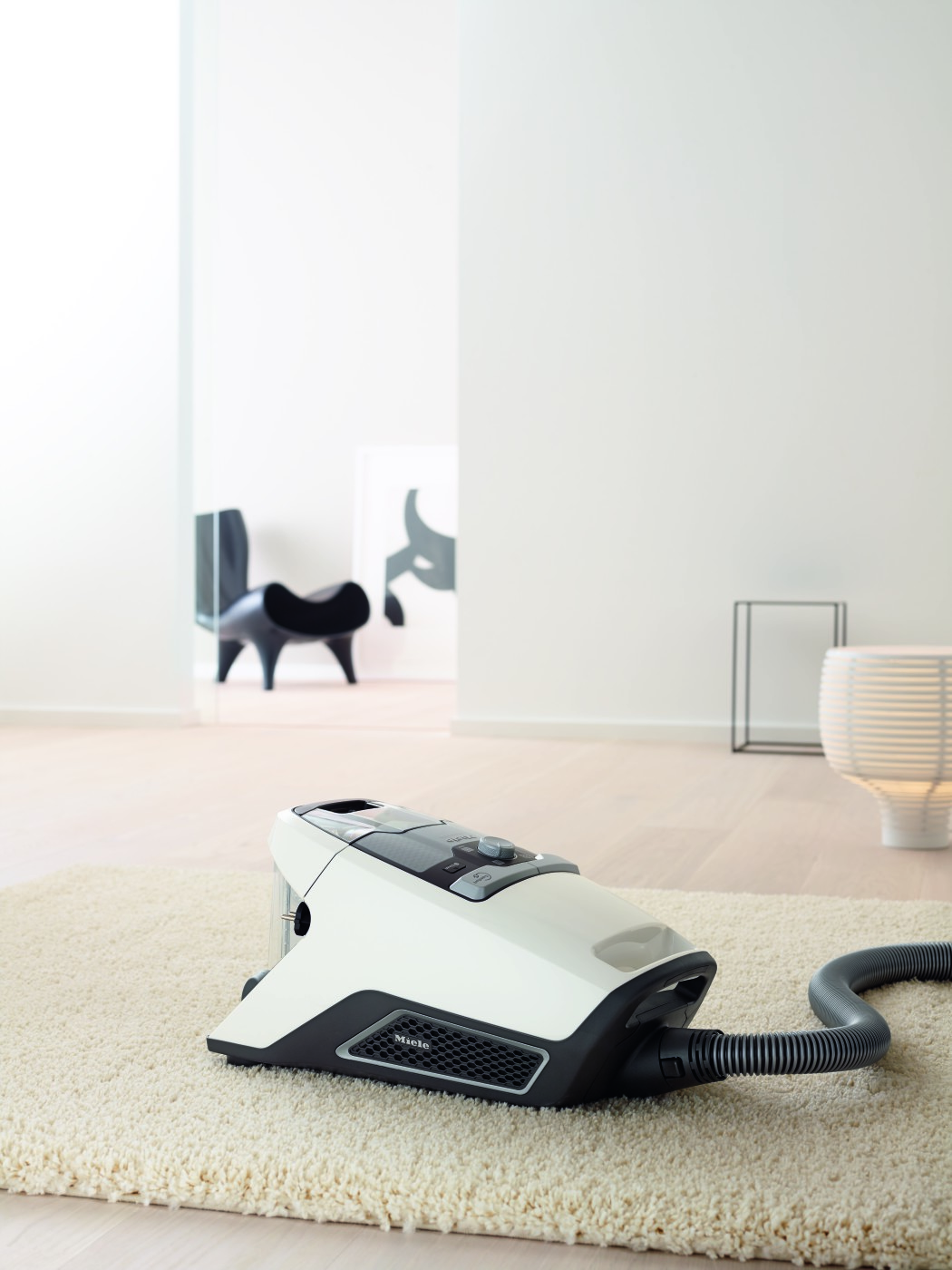 White vacuum cleaner on a beige carpet. A black chair and a lamp are in the background.