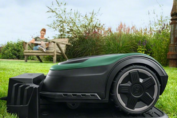 Robotfűnyíró a füvön. Egy nő a háttérben egy padon ülve olvas. Zöld és fekete.