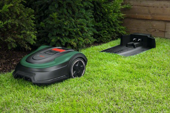 Egy robotfűnyíró a füvön, a töltőállomás mellett. Zöld fű és sövény a háttérben.