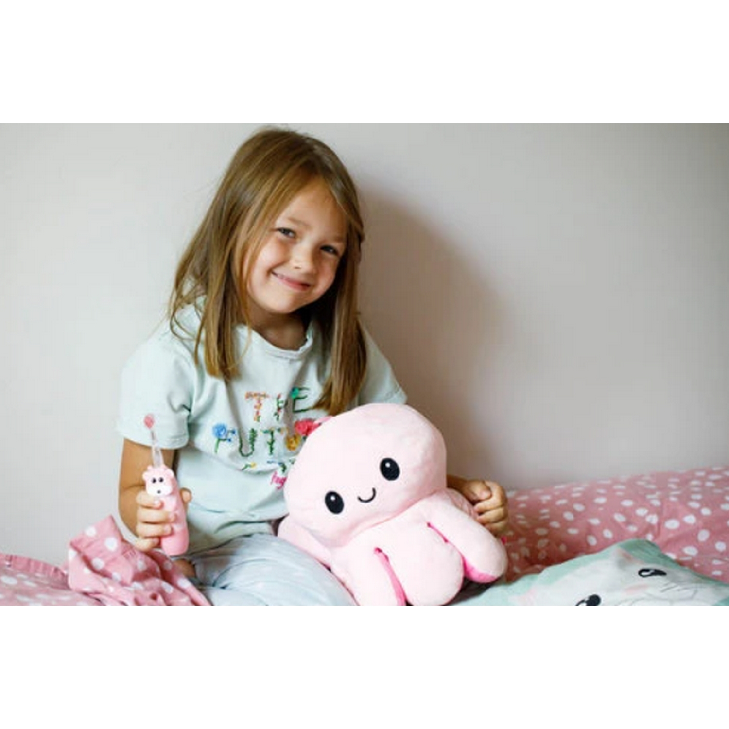 Una niña sonríe, sosteniendo un peluche de pulpo rosa y un cepillo de dientes rosa y blanco.