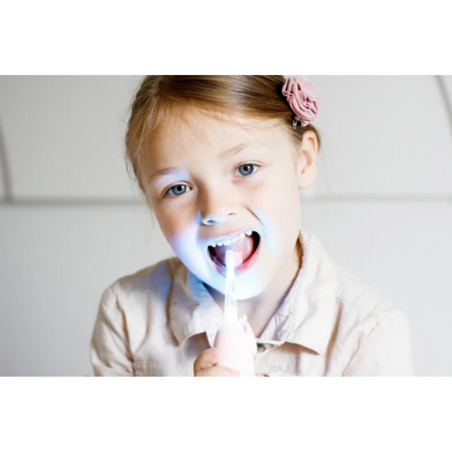 Una niña con una flor rosa en el pelo usa un cepillo de dientes con luz azul. Tiene la boca abierta.