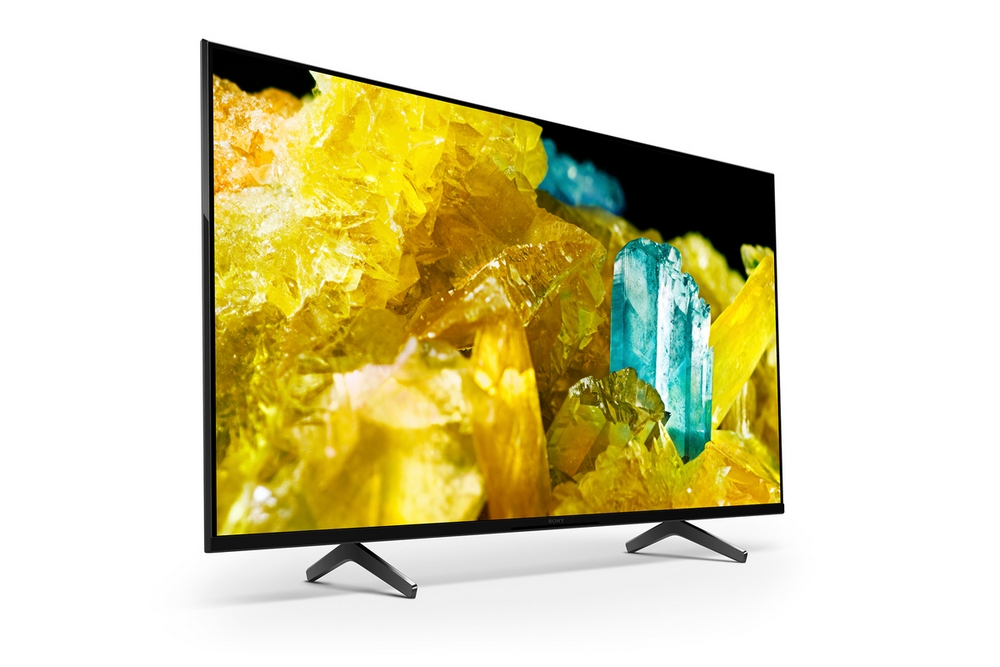 Un televisor grande muestra arte abstracto amarillo y azul. Se apoya en patas negras sobre un fondo blanco.