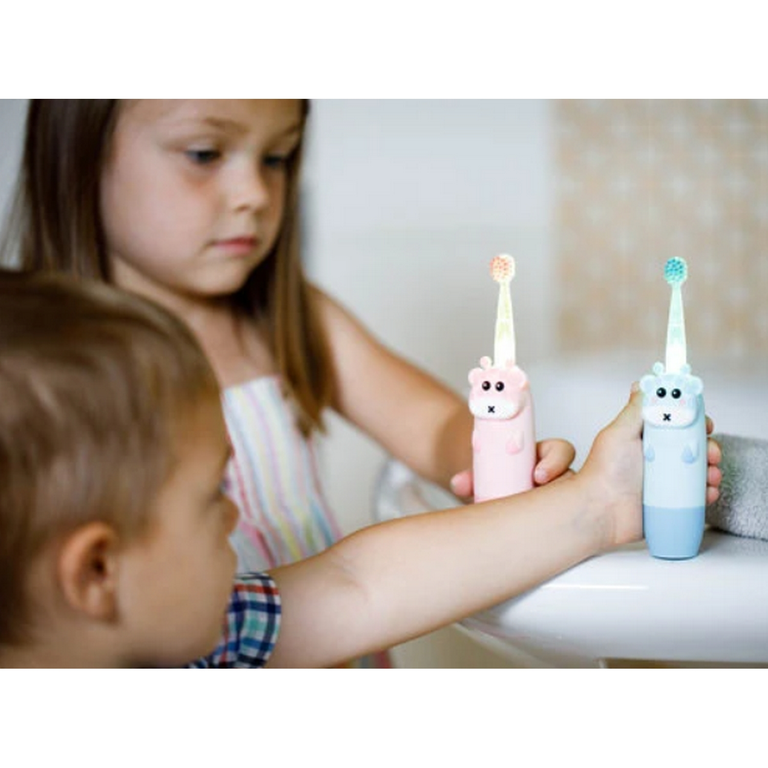 Dos niños con cepillos de dientes con forma de hámster. Uno rosa, uno azul, en un lavabo blanco.