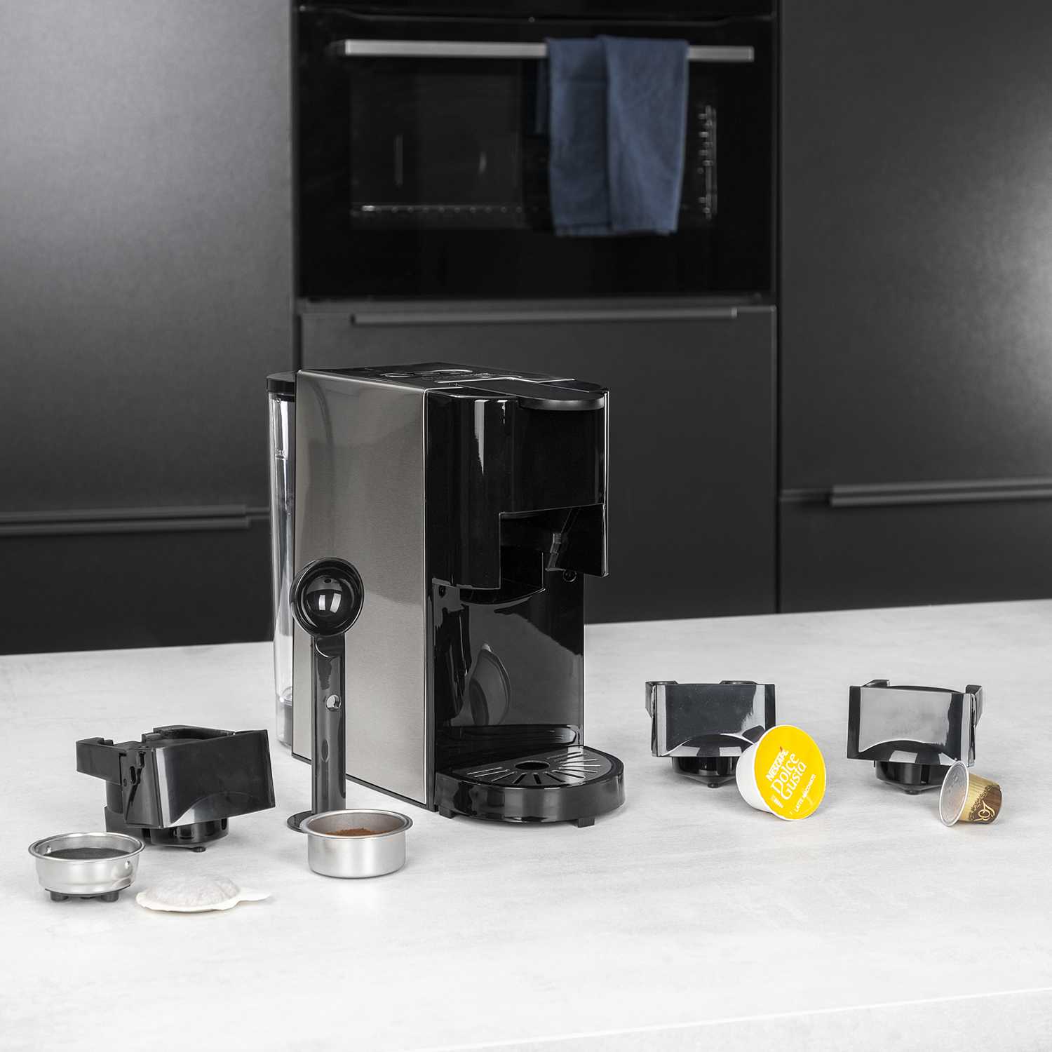 Een zilver-zwarte koffiemachine met koffiepads, een watertank en accessoires op een wit aanrecht.