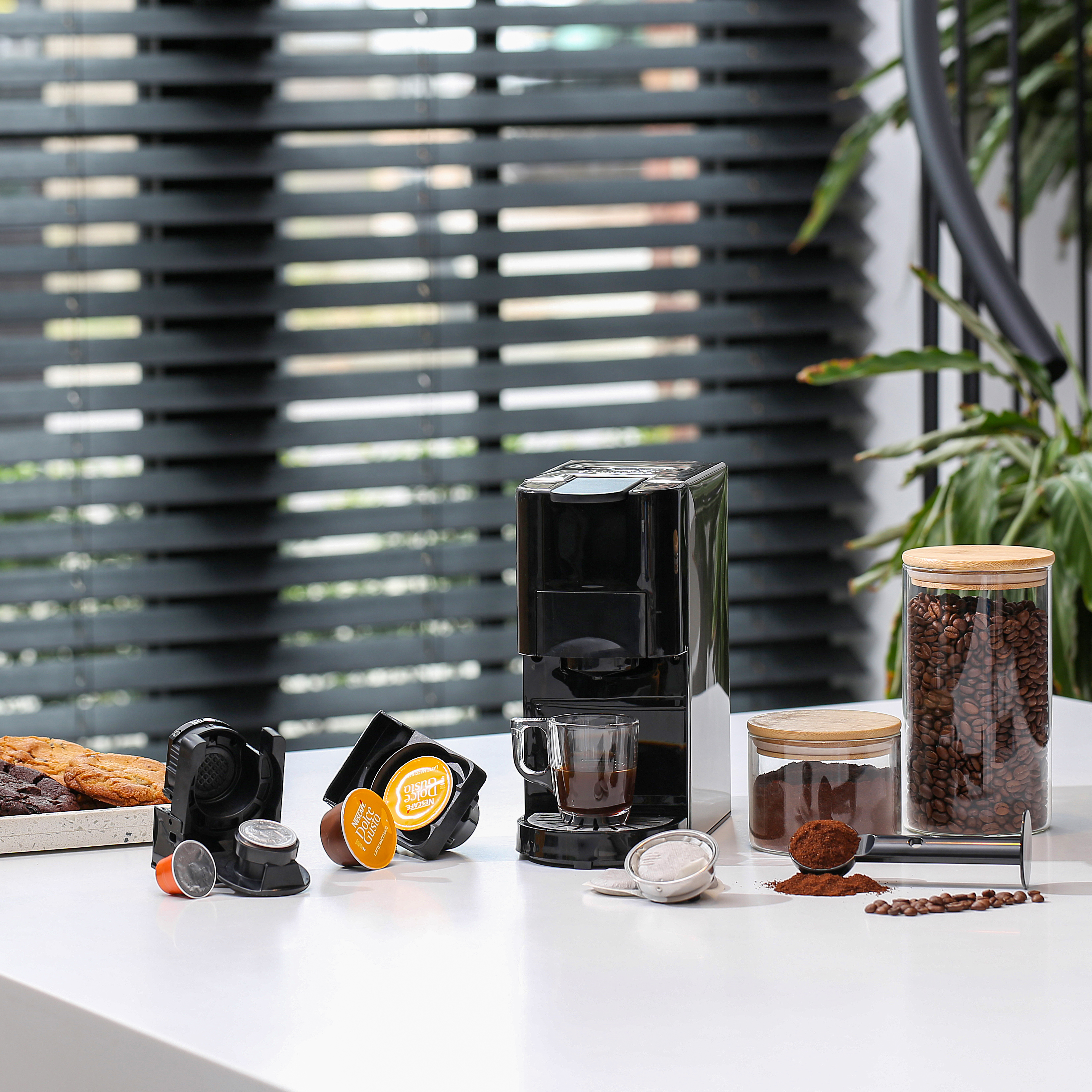 Zwarte espressomachine met kopjes, koffiecapsules, bonen en accessoires op een wit oppervlak.