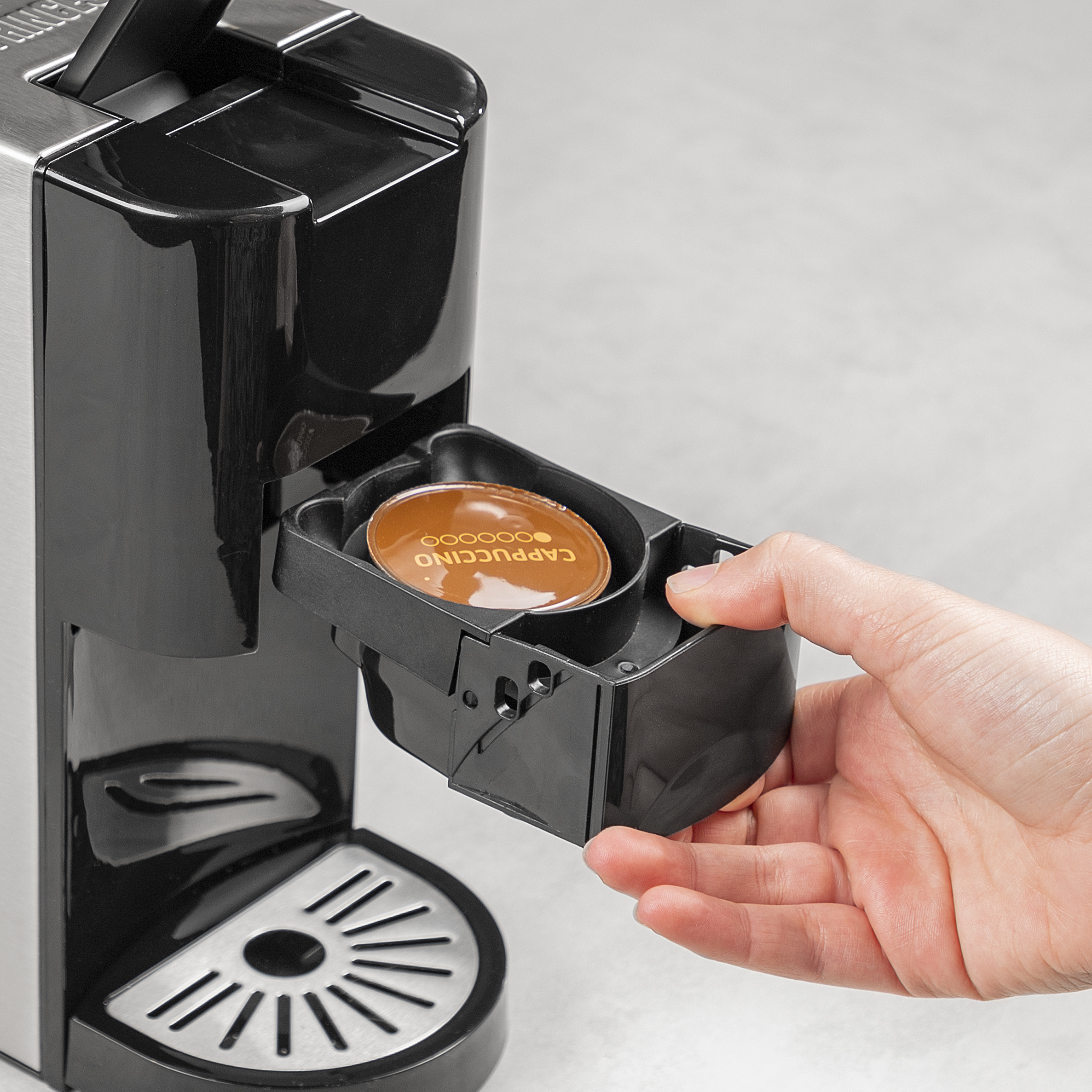 Persoon voegt een 'Cappuccino' koffiepad toe aan een zwart-zilveren koffiemachine.