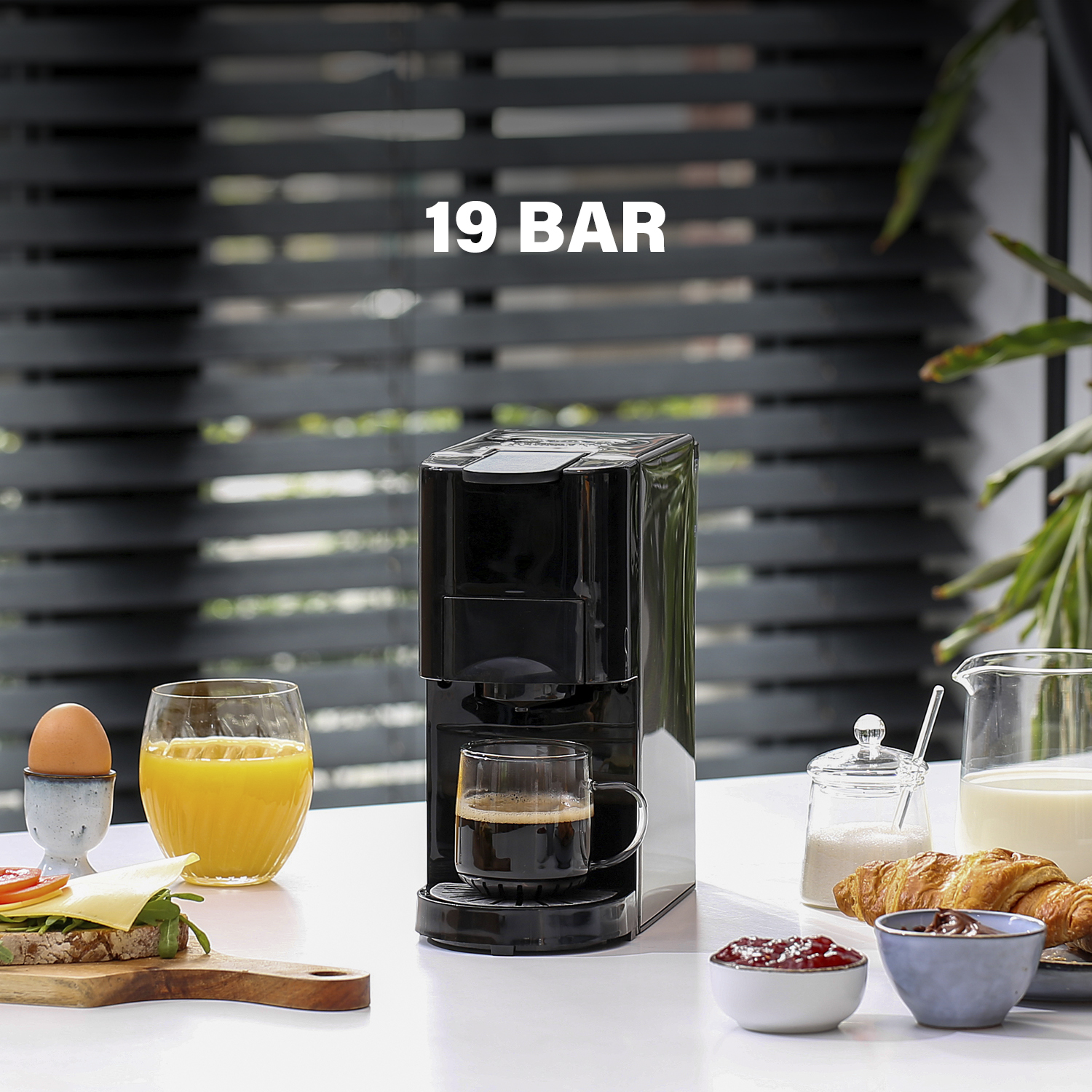 Zwart koffiezetapparaat met een glas koffie en ontbijt op een tafel.