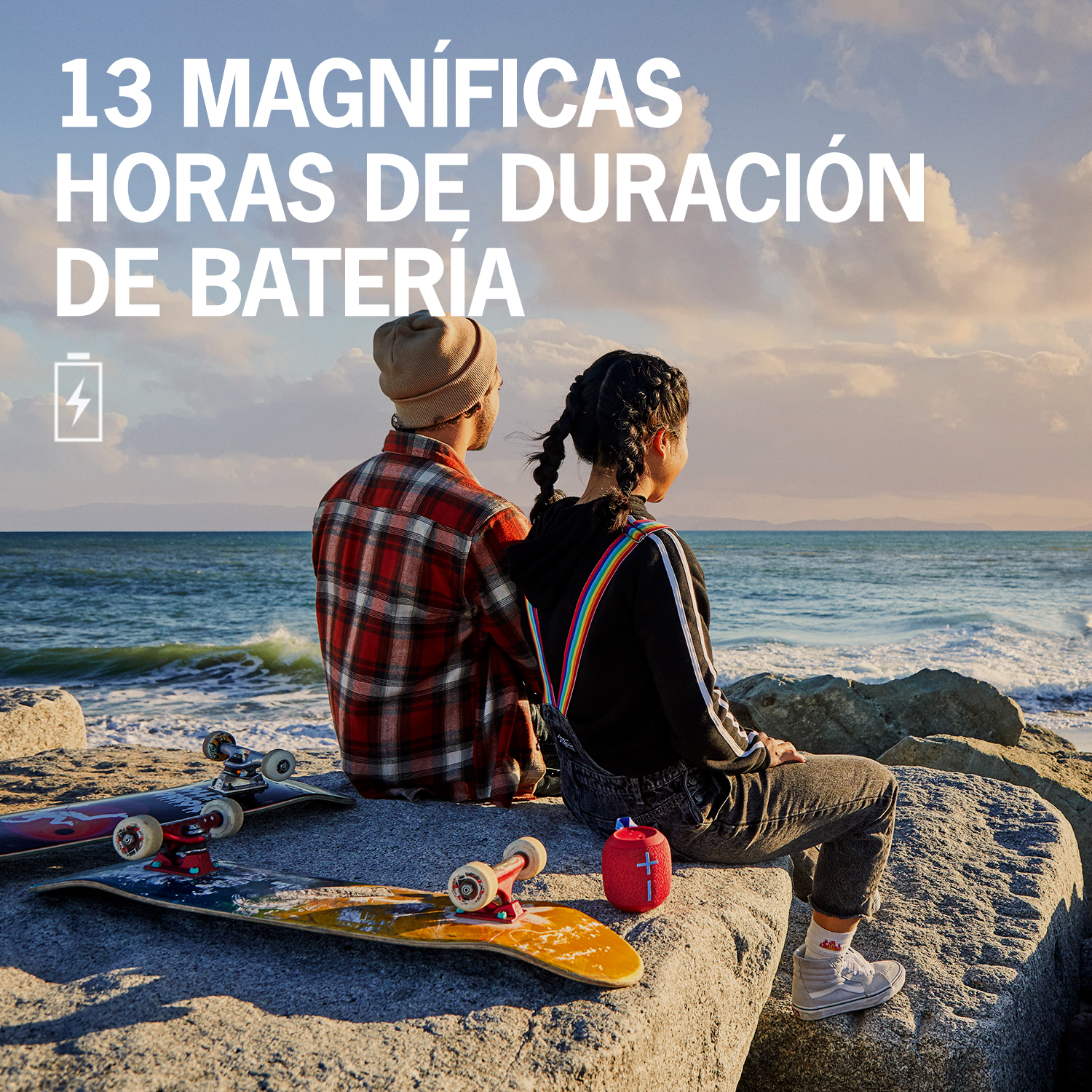 Dos personas sentadas en rocas junto al mar, con monopatines y un altavoz.