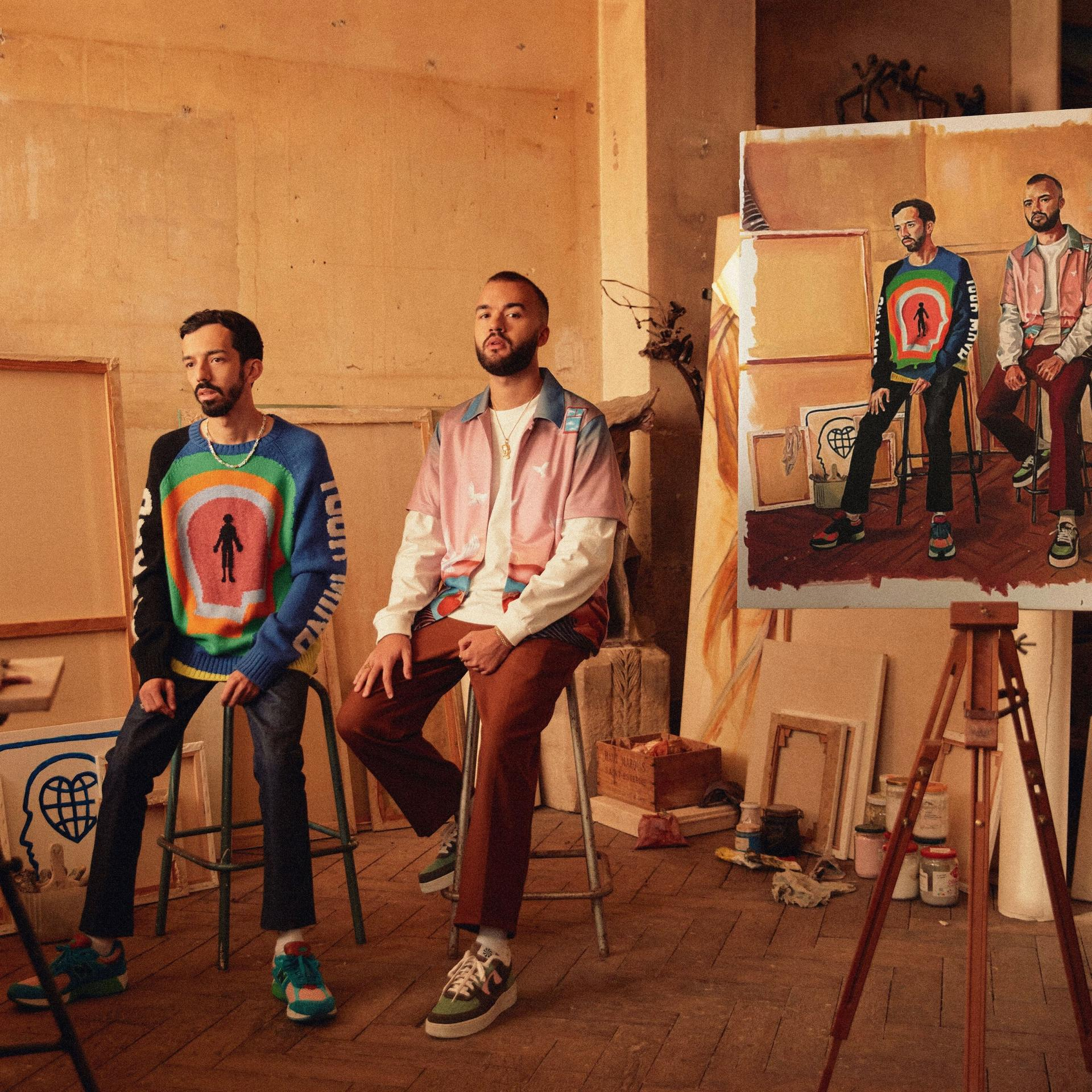 Two men in an art studio, one seated, another standing near a painting.