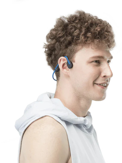 Un jeune homme aux cheveux bouclés portant un sweat à capuche blanc sourit tout en portant des écouteurs bleus et noirs.