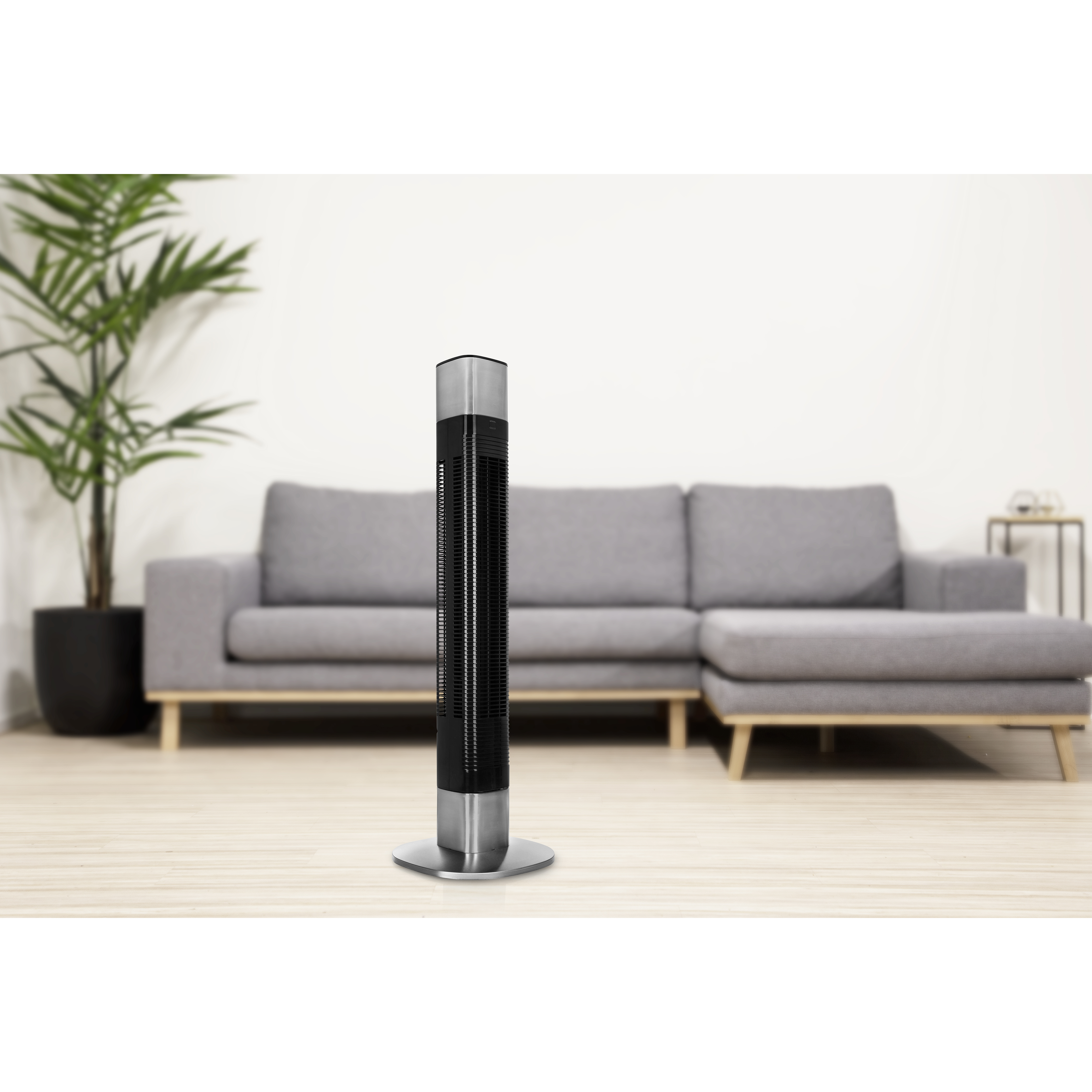 Een zwarte en zilveren torenventilator voor een grijze bank. Een plant staat links.