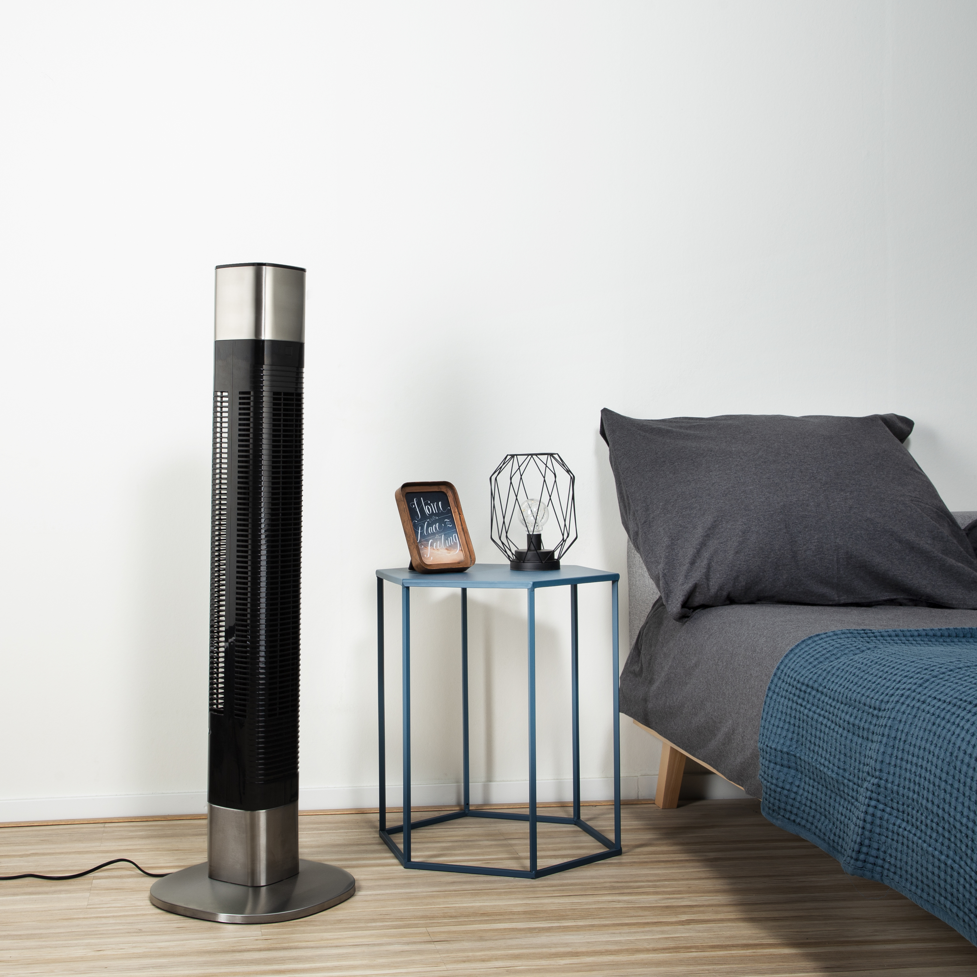 Zwarte en zilveren torenventilator naast een blauwe tafel met een fotolijst en een lamp.