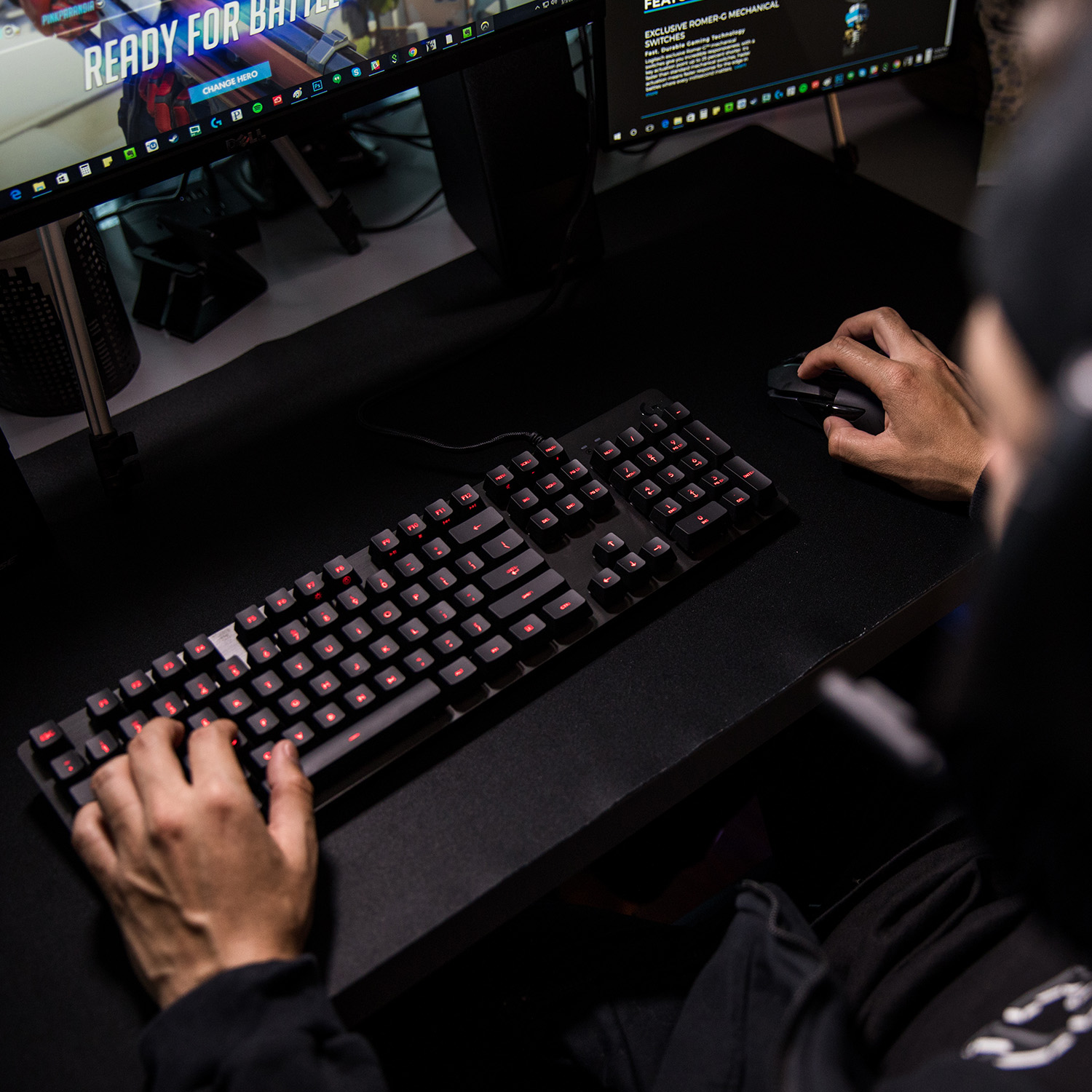 Manos en teclado con teclas retroiluminadas en rojo. Ratón a la derecha, monitor al fondo.