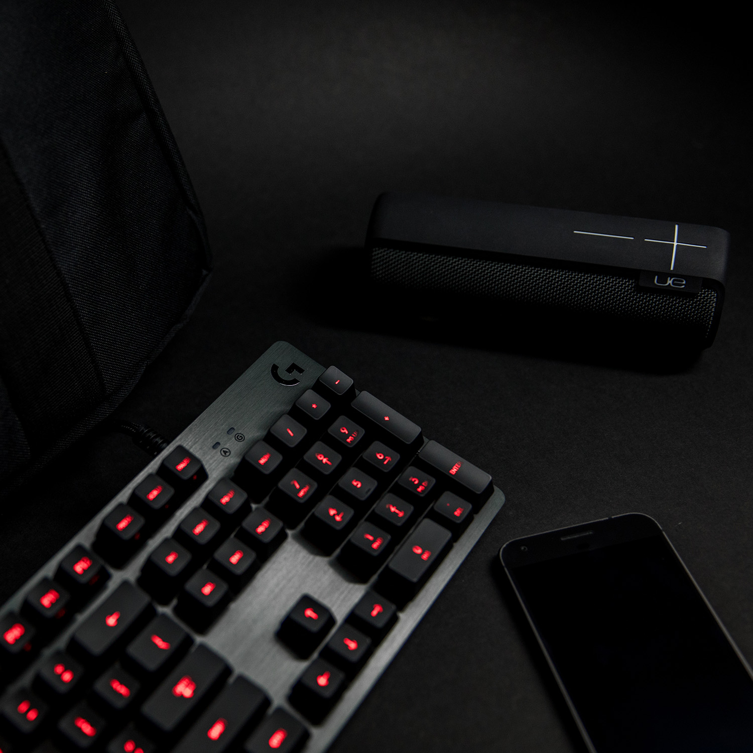 Teclado negro con teclas retroiluminadas en rojo, un altavoz negro y un smartphone negro.