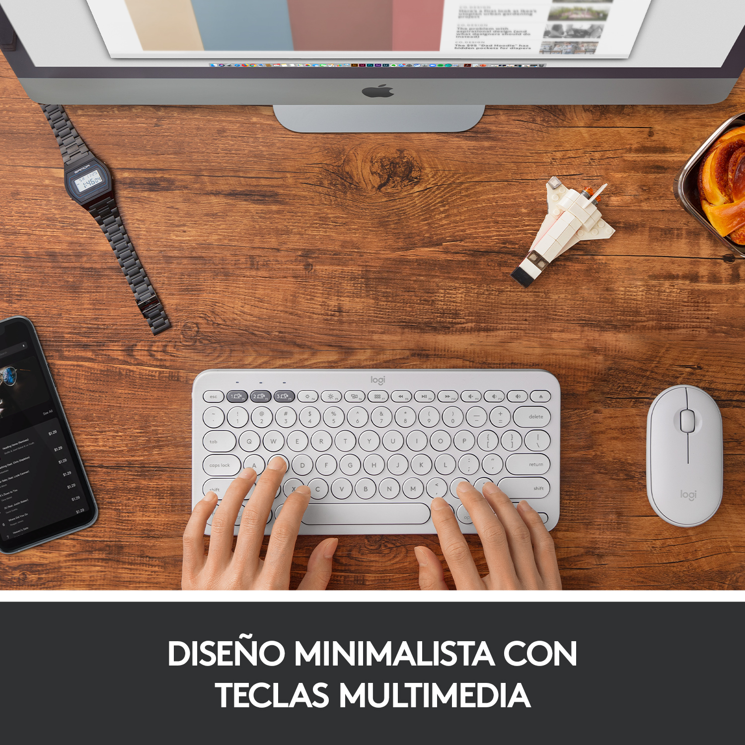Manos escribiendo en teclado blanco, teléfono, reloj, ratón y ordenador en mesa de madera. Diseño minimalista.