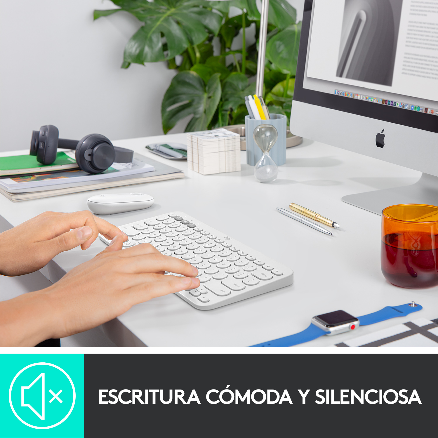Una persona escribiendo en un teclado blanco. Monitor de Apple, auriculares y reloj en el escritorio.