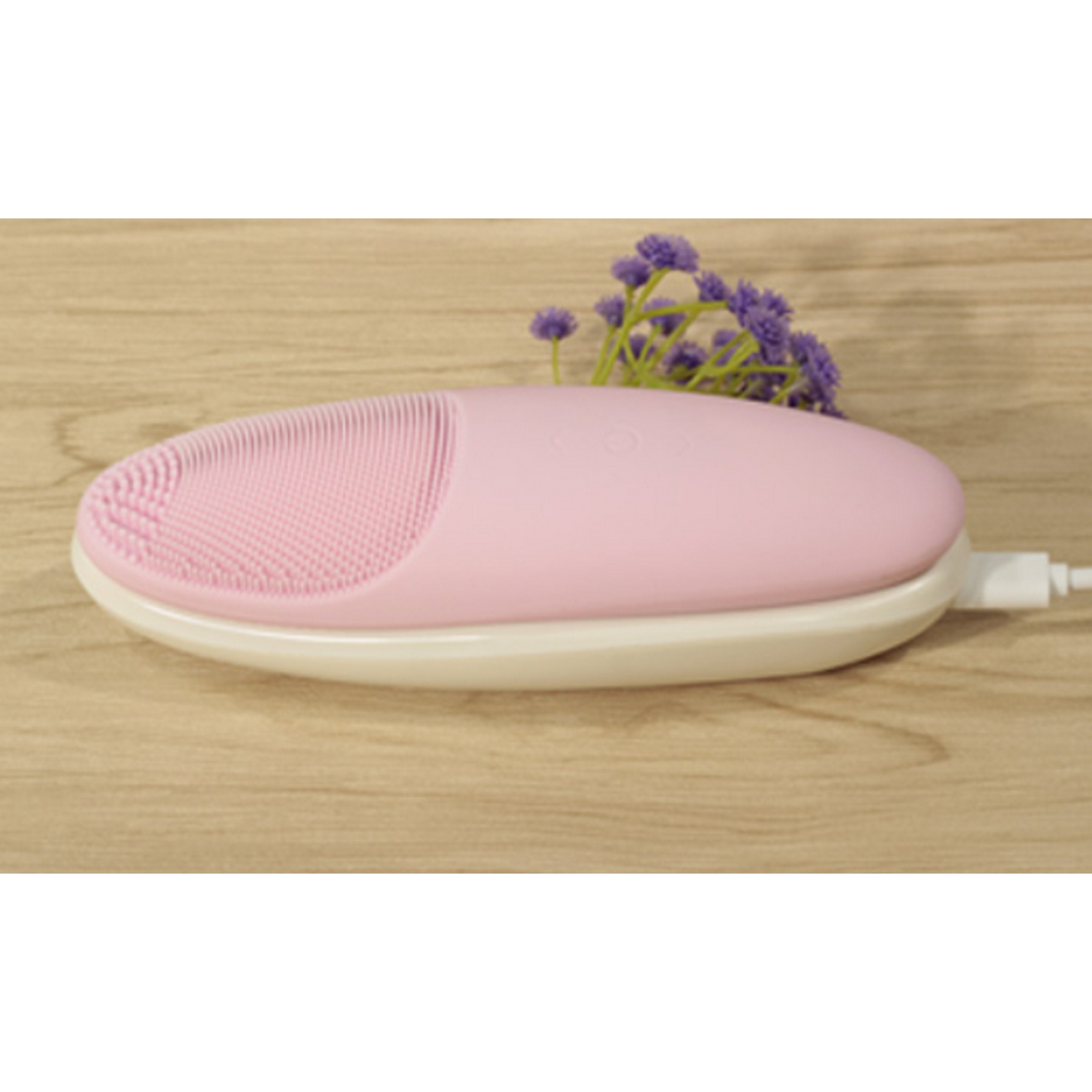 Cepillo de limpieza facial rosa sobre base blanca, con flores moradas en superficie de madera.