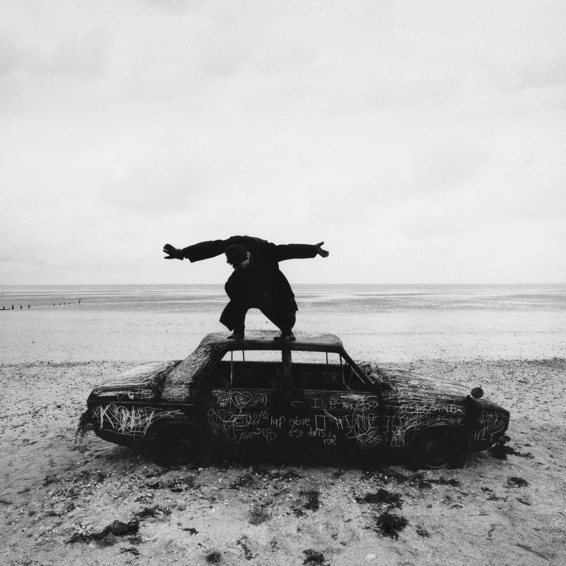 Een persoon die op een auto op een strand staat. De auto is beklad. Bewolkte lucht.