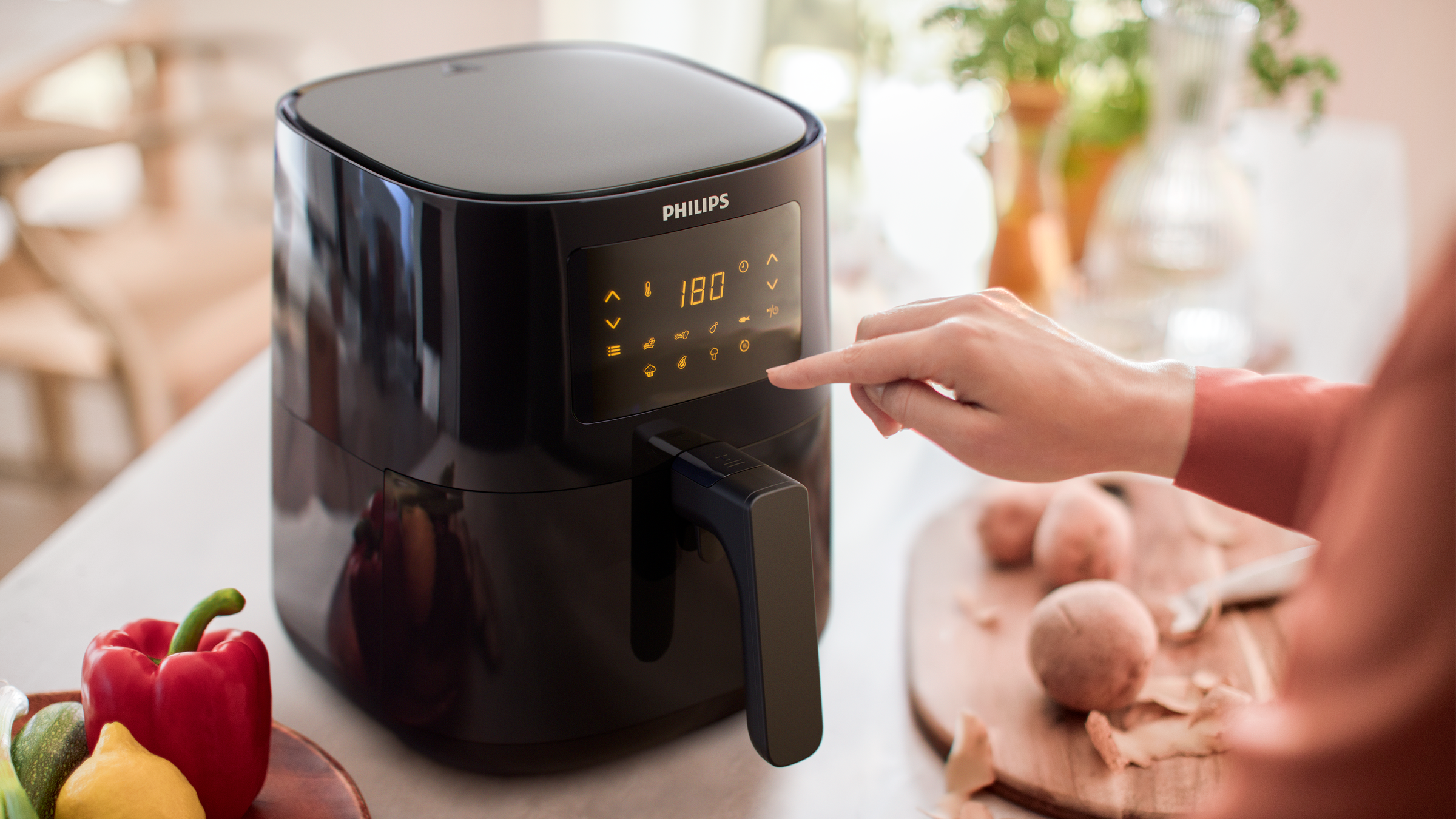 Black air fryer with a woman's hand touching the digital display. Vegetables and a cutting board are visible.