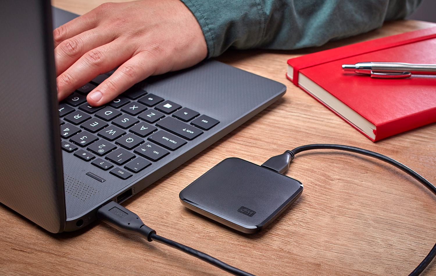 A hand typing on a laptop keyboard. A black external hard drive is connected. A red notebook sits beside it.