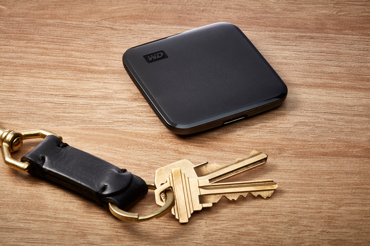 Black external hard drive next to keys and a keychain on a wooden surface.