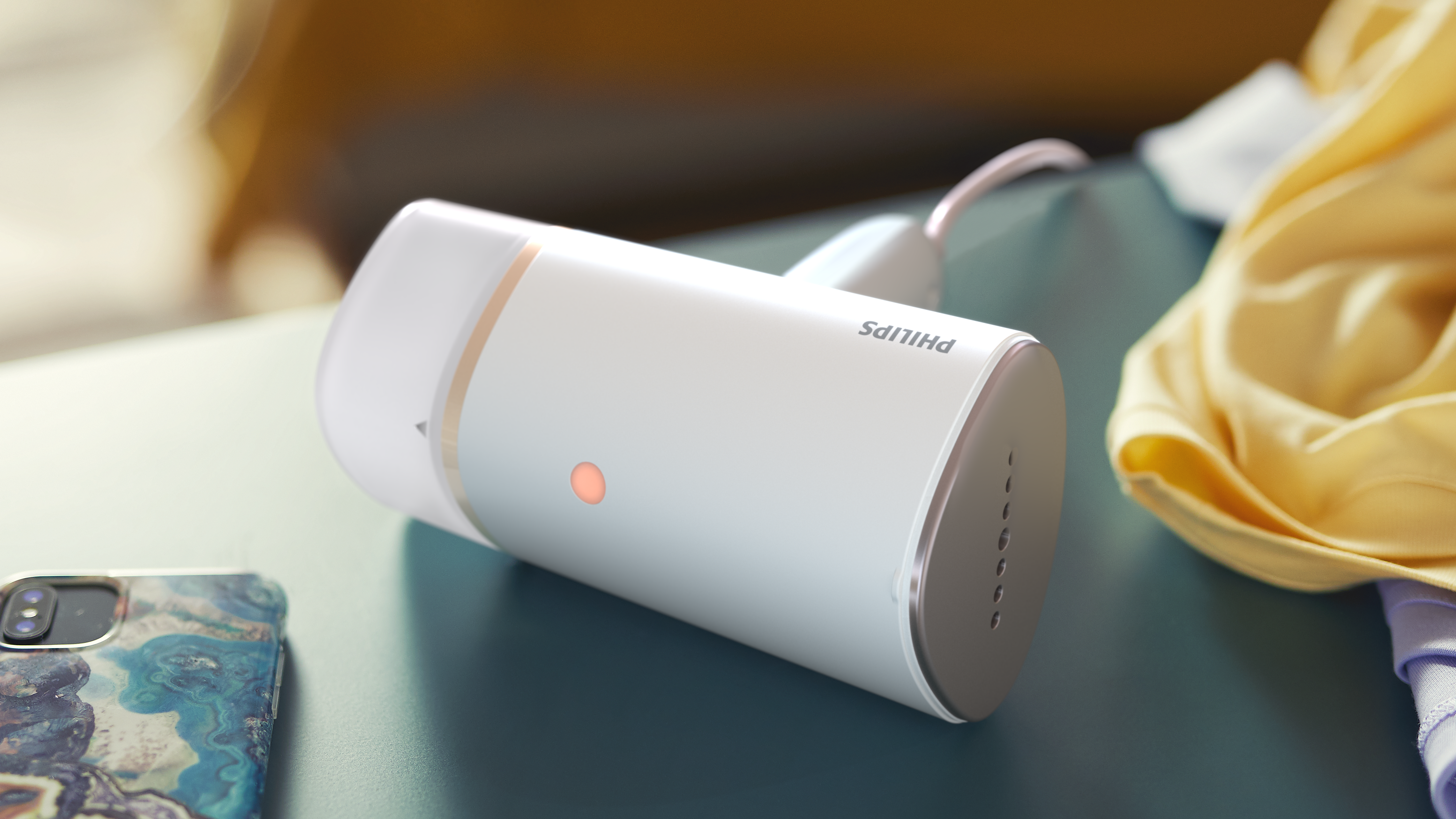 Una plancha de vapor Philips blanca con un botón iluminado está sobre una superficie verde azulado, con un teléfono y tela amarilla cerca.