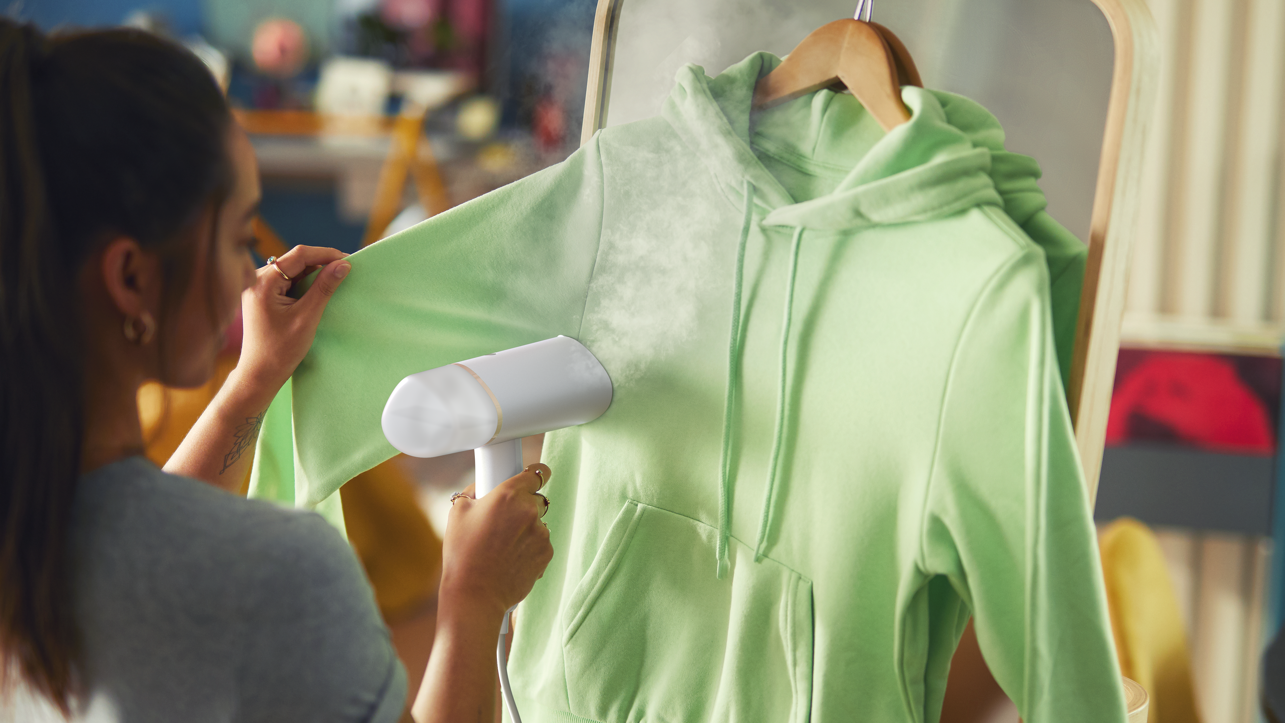 Una mujer plancha con vapor una sudadera con capucha verde con un vaporizador blanco. La sudadera cuelga de una percha de madera. El vapor sale de la tela.