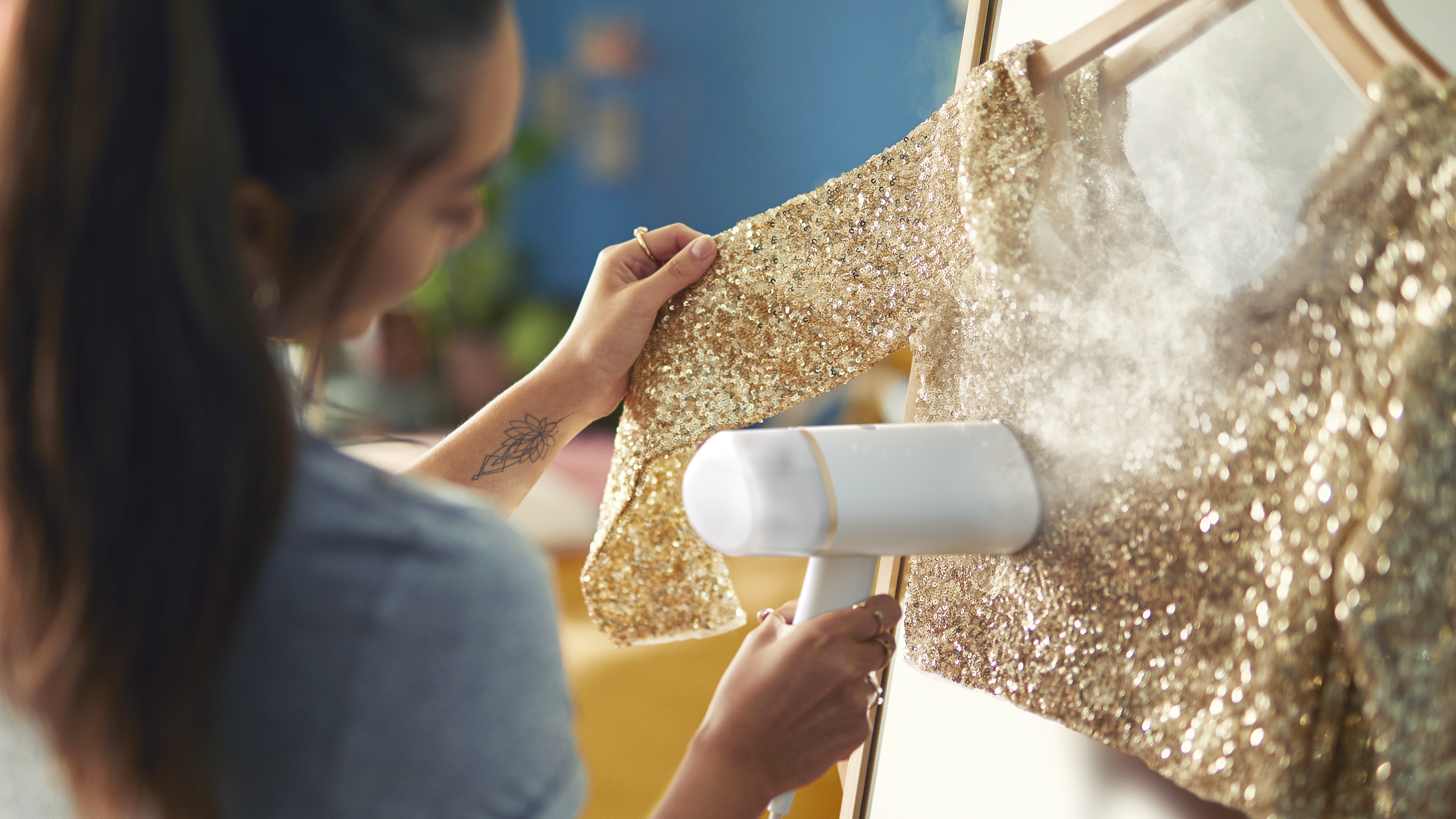 Una mujer plancha un vestido dorado con lentejuelas con un vaporizador de mano. El vapor ondea. El vestido cuelga de una percha de madera.
