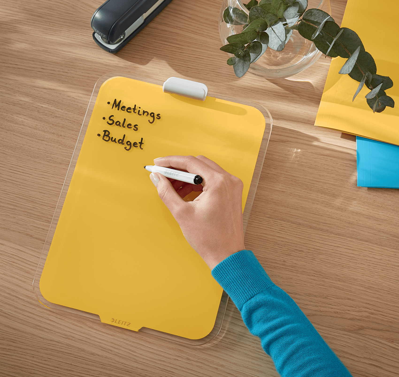 Egy kéz ír egy sárga irattáblára tollal. A táblán a következő szöveg szerepel: Meetings, Sales, Budget.